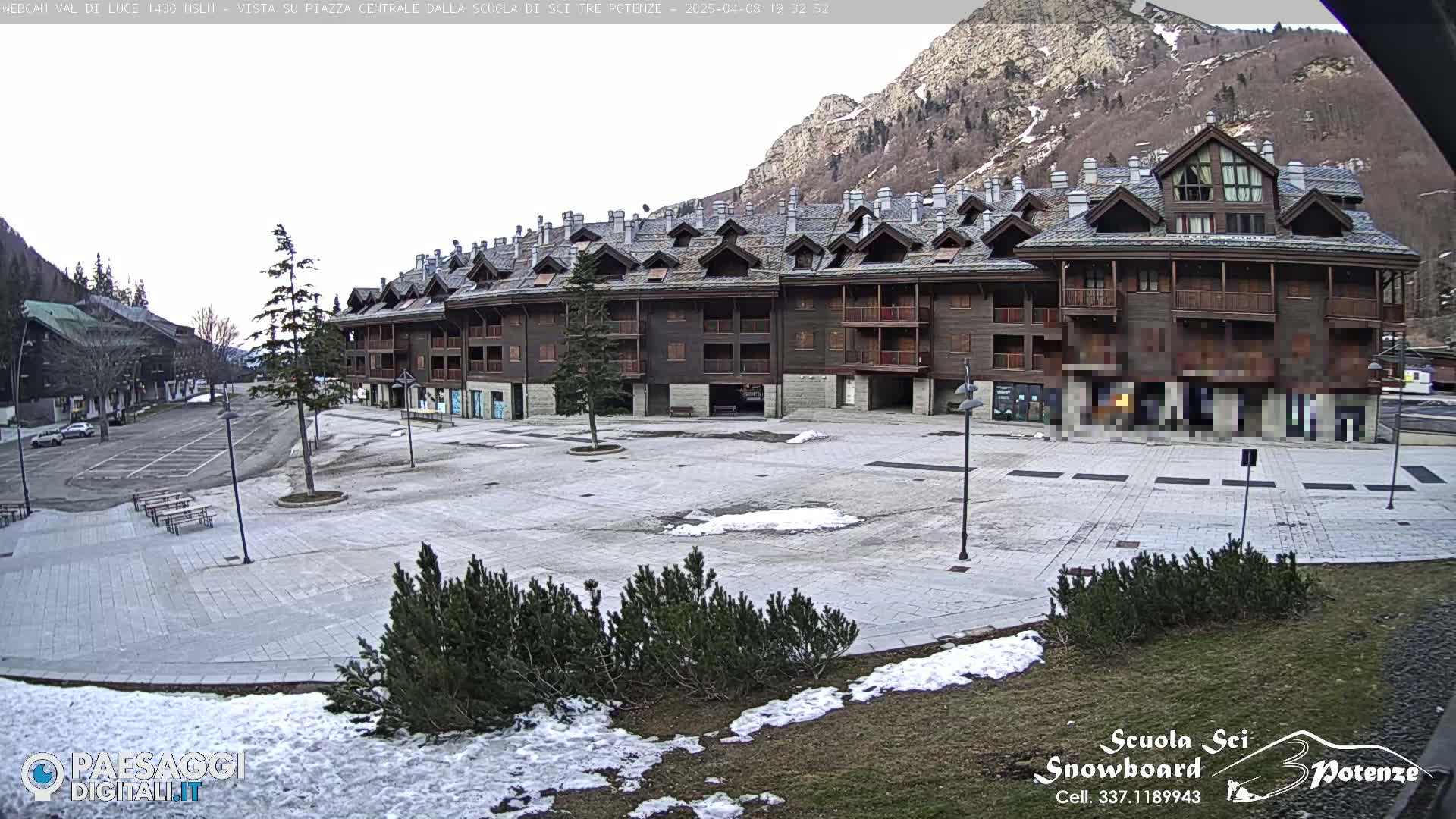 A large, empty plaza with a long, multi-story building behind it, surrounded by snow patches and evergreens, under an overcast sky.