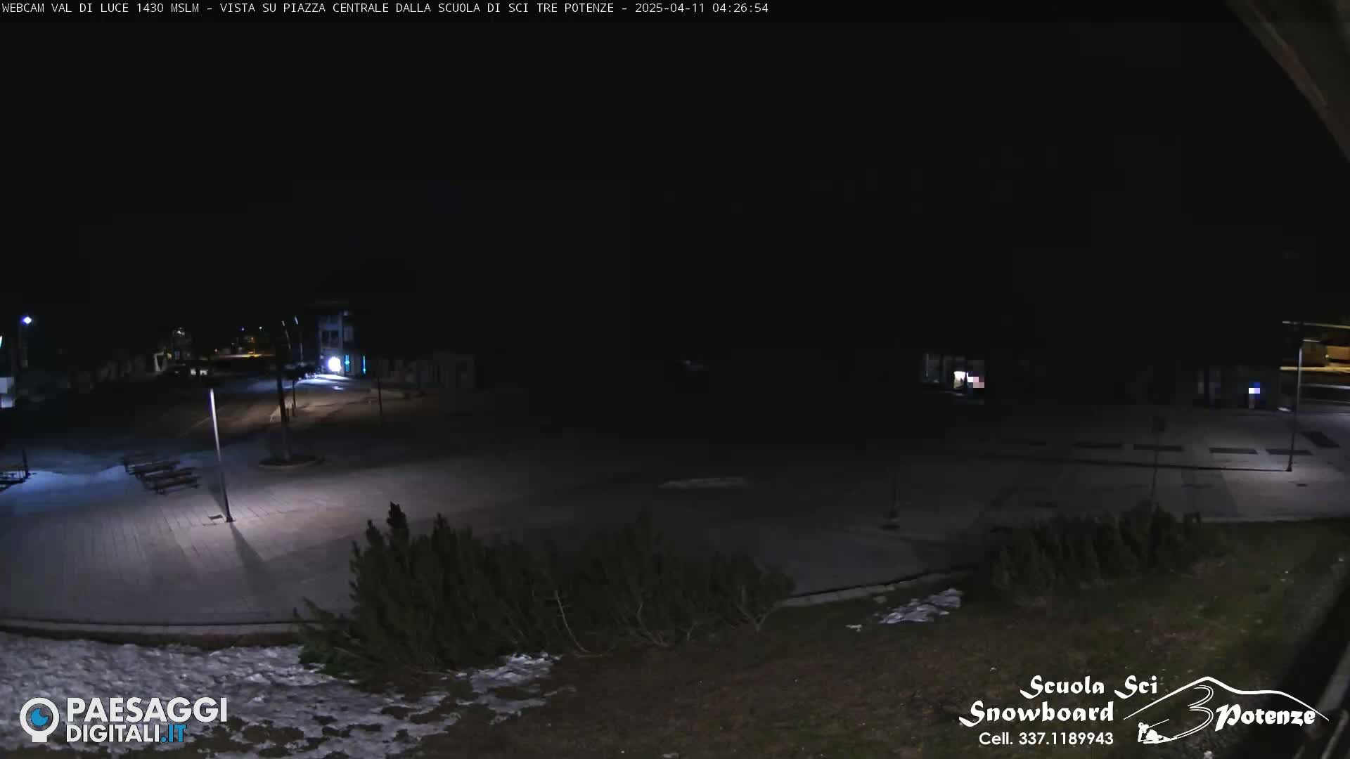 A nighttime view of a snow-dusted, empty plaza with some sparse shrubs and low-lying buildings under a dark sky.