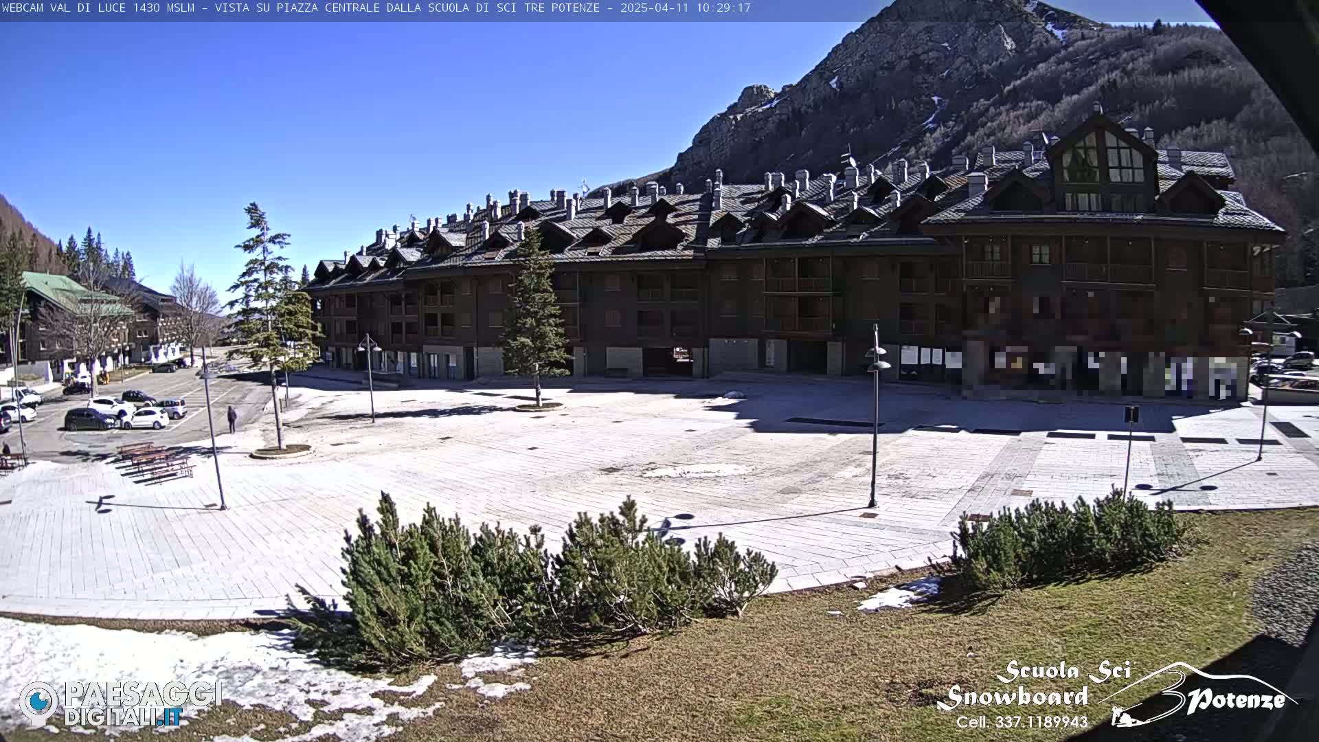 A large, paved plaza sits in front of a multi-story building, under a clear blue sky, with some snow patches visible on the ground and mountains in the background.