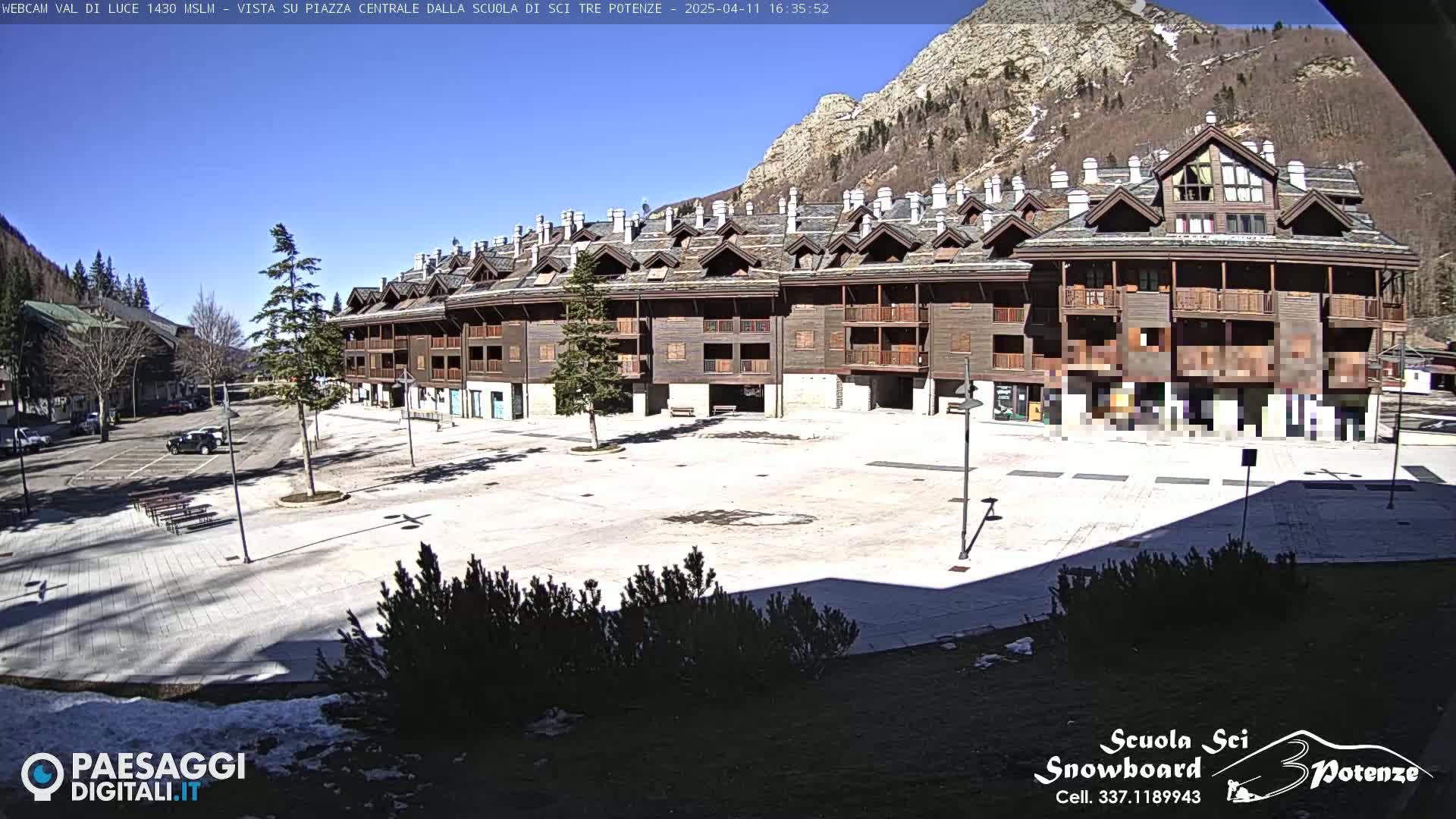 A large, empty plaza is situated in front of a long, multi-story brown building nestled against a mountainside under a clear blue sky.