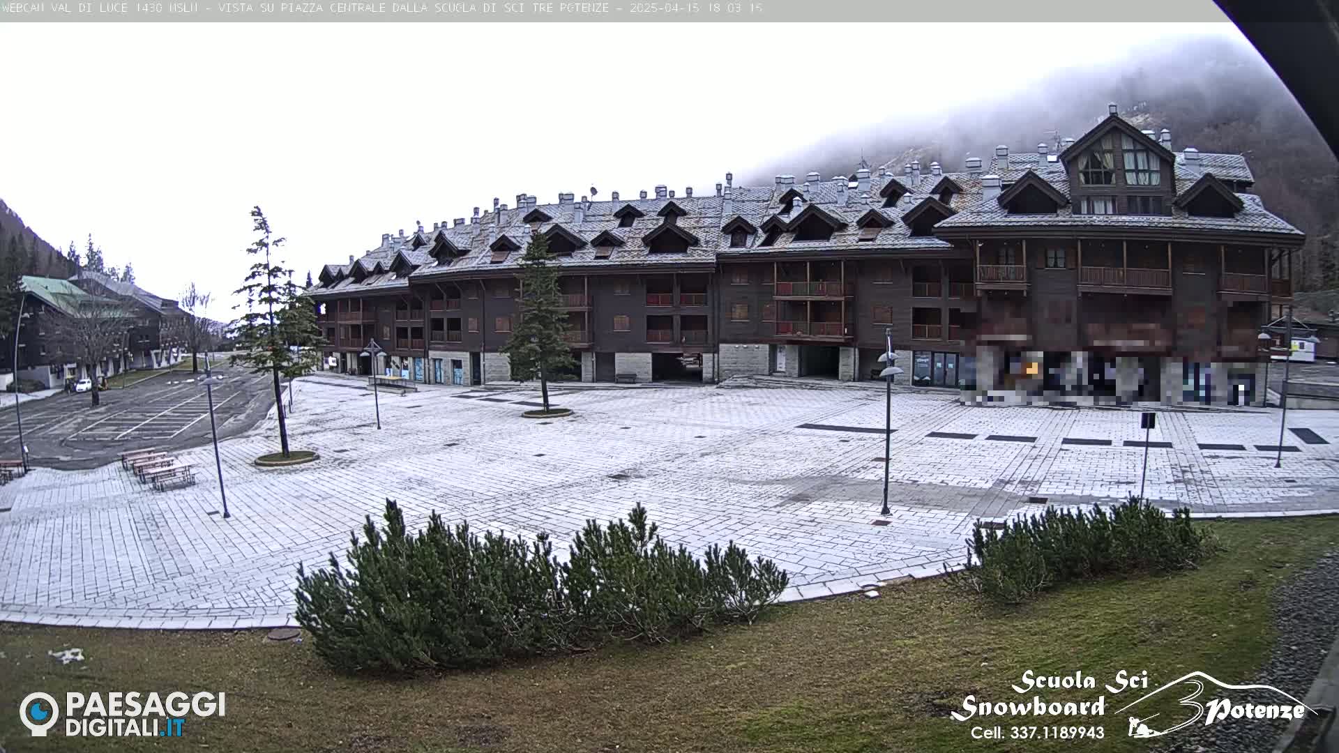 A snow-dusted stone plaza sits in front of a large, multi-story dark brown building, with a mountain shrouded in fog in the background.