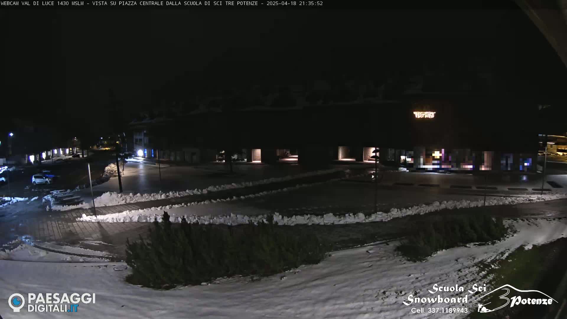 A nighttime view of a snow-dusted town square with low-level lighting, a few parked cars, and small shrubs.