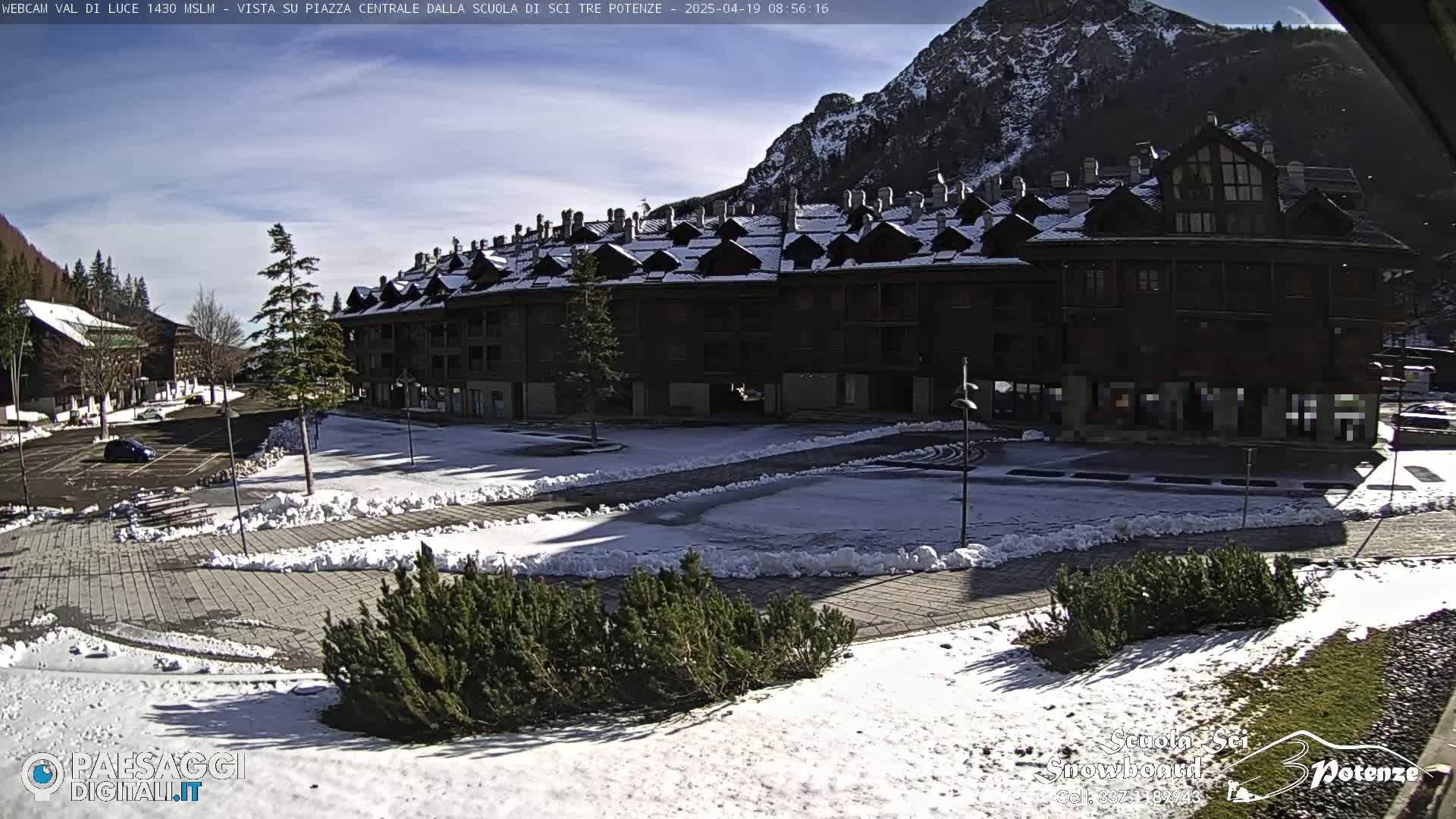 A snow-dusted plaza with a large, multi-story building and mountains in the background under a mostly sunny sky.