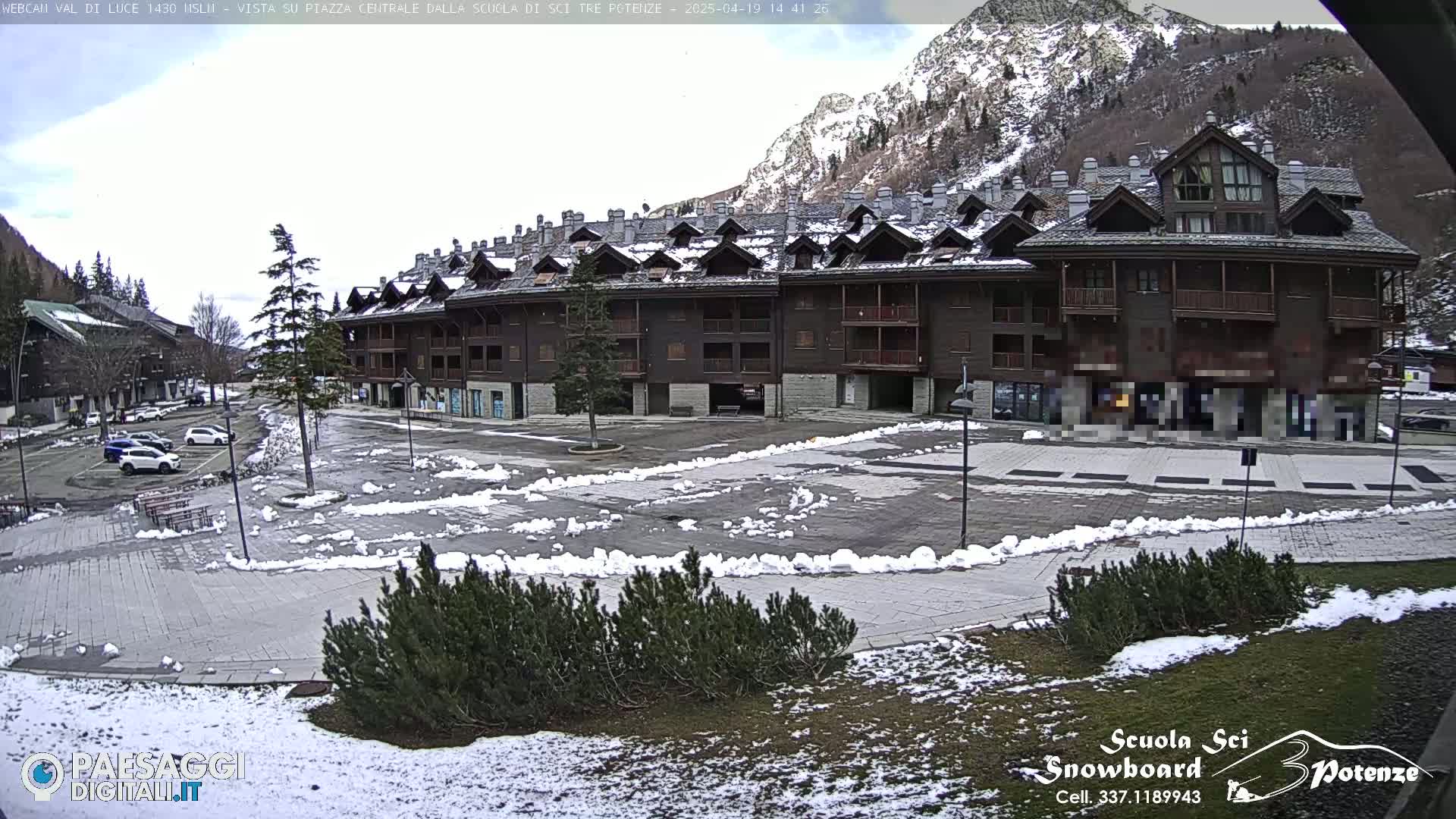 A snow-dusted plaza with a large, dark brown multi-story building sits in front of a mountain range under a mostly clear sky.