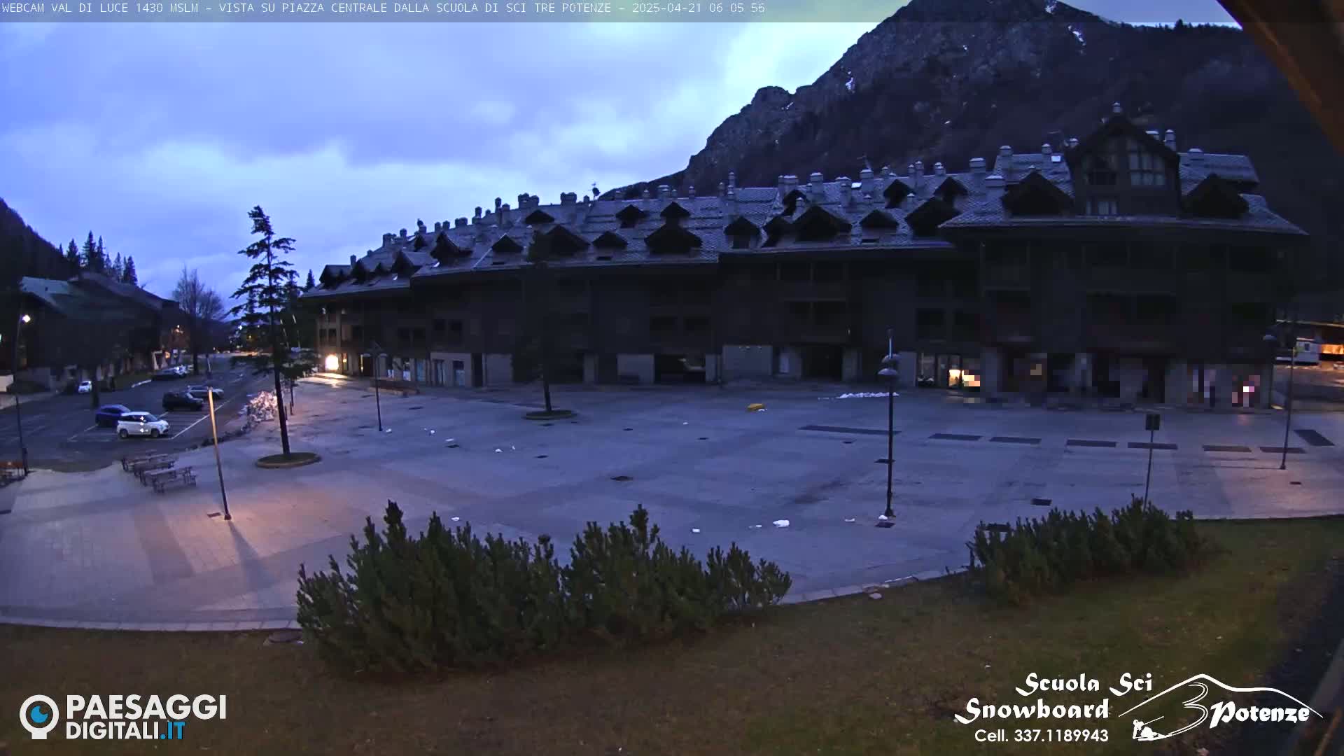A mostly empty town square sits before a large, dark-colored building at dusk, with mountains visible in the background under a cloudy sky.