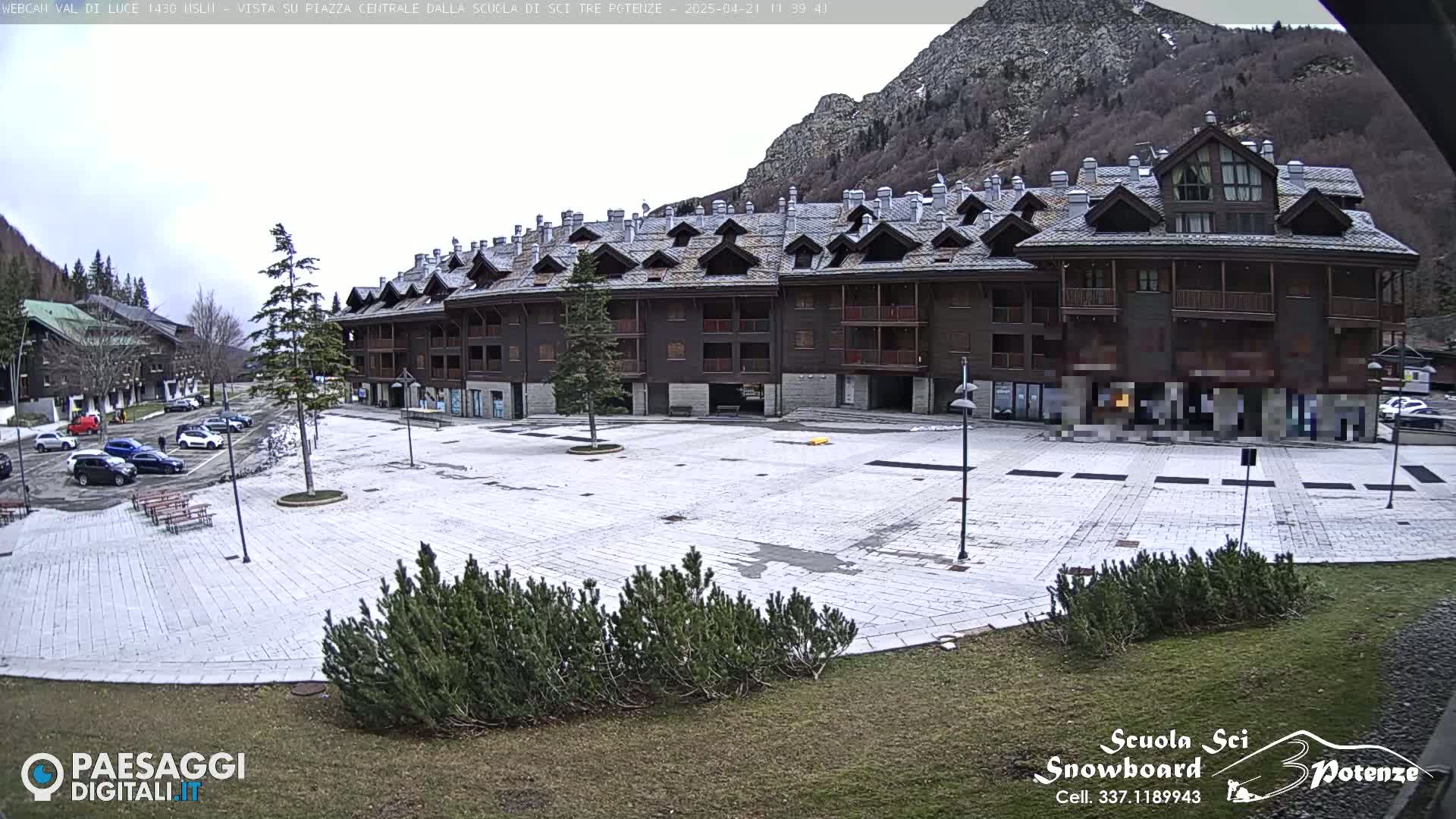 A large, empty paved plaza sits in front of a multi-story brown building, with mountains and overcast skies in the background.