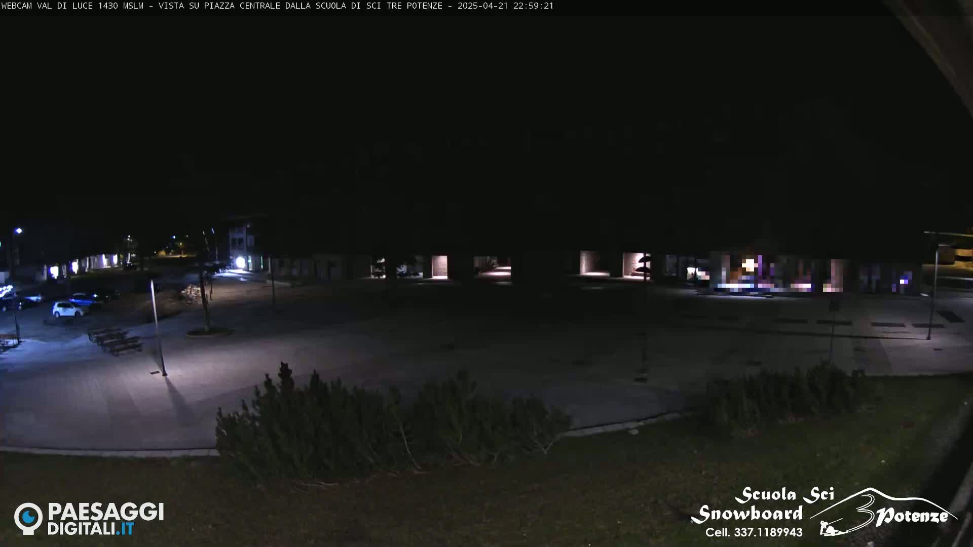 A nighttime, empty plaza is shown, illuminated by artificial lights, with a few cars parked nearby.