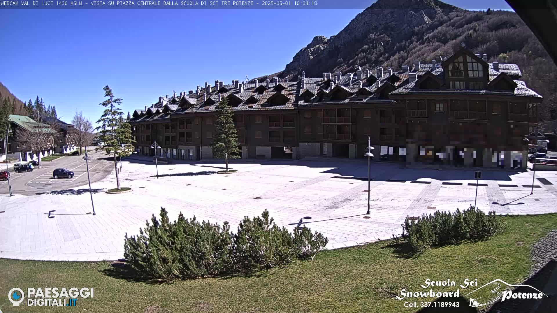 A large, empty plaza paved in light-colored stone is situated in front of a multi-story building in a mountainous area under a clear, sunny sky.