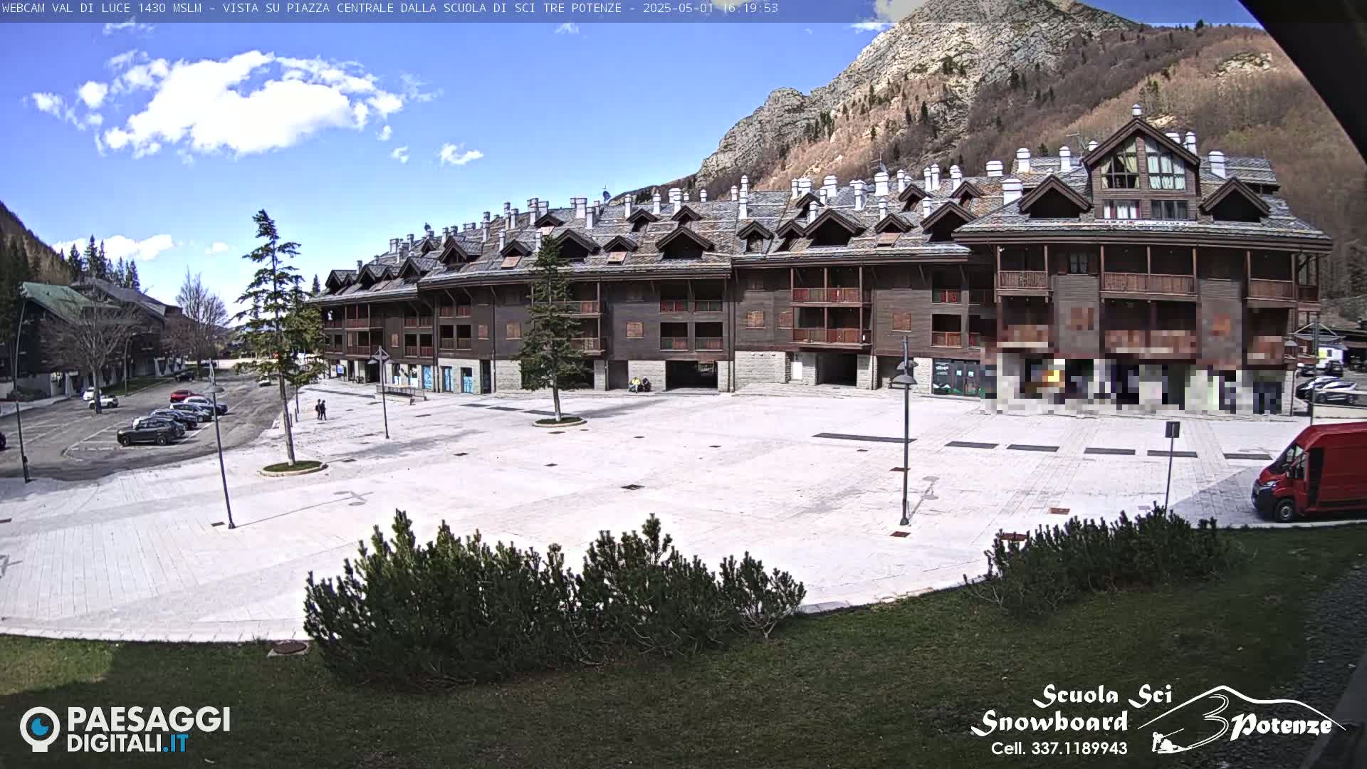 A large, empty plaza is in front of a multi-story, dark brown building with balconies, under a partly cloudy sky in a mountainous region.