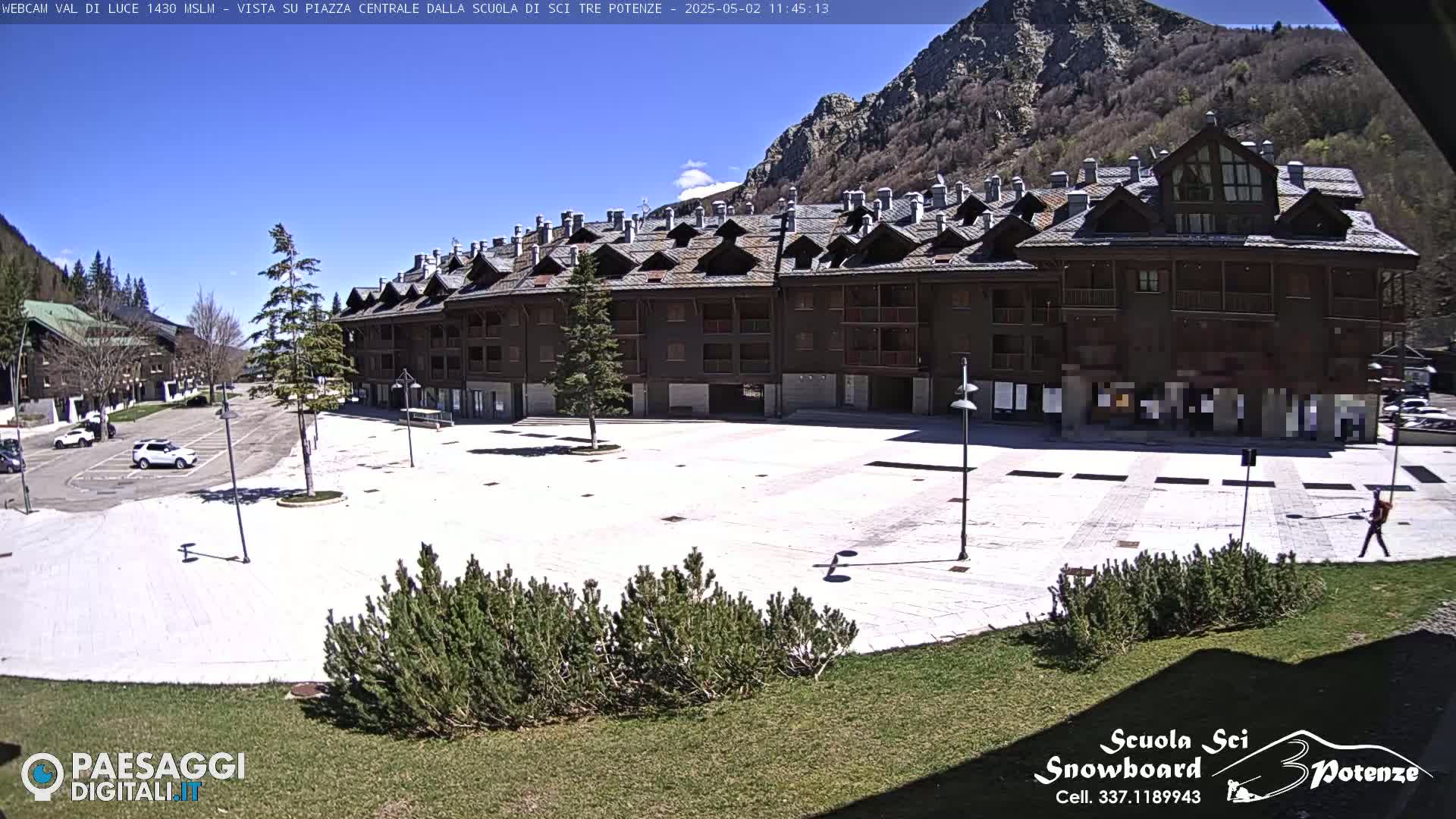 A large, light-colored plaza sits in front of a multi-story brown building, backed by mountains under a clear blue sky.