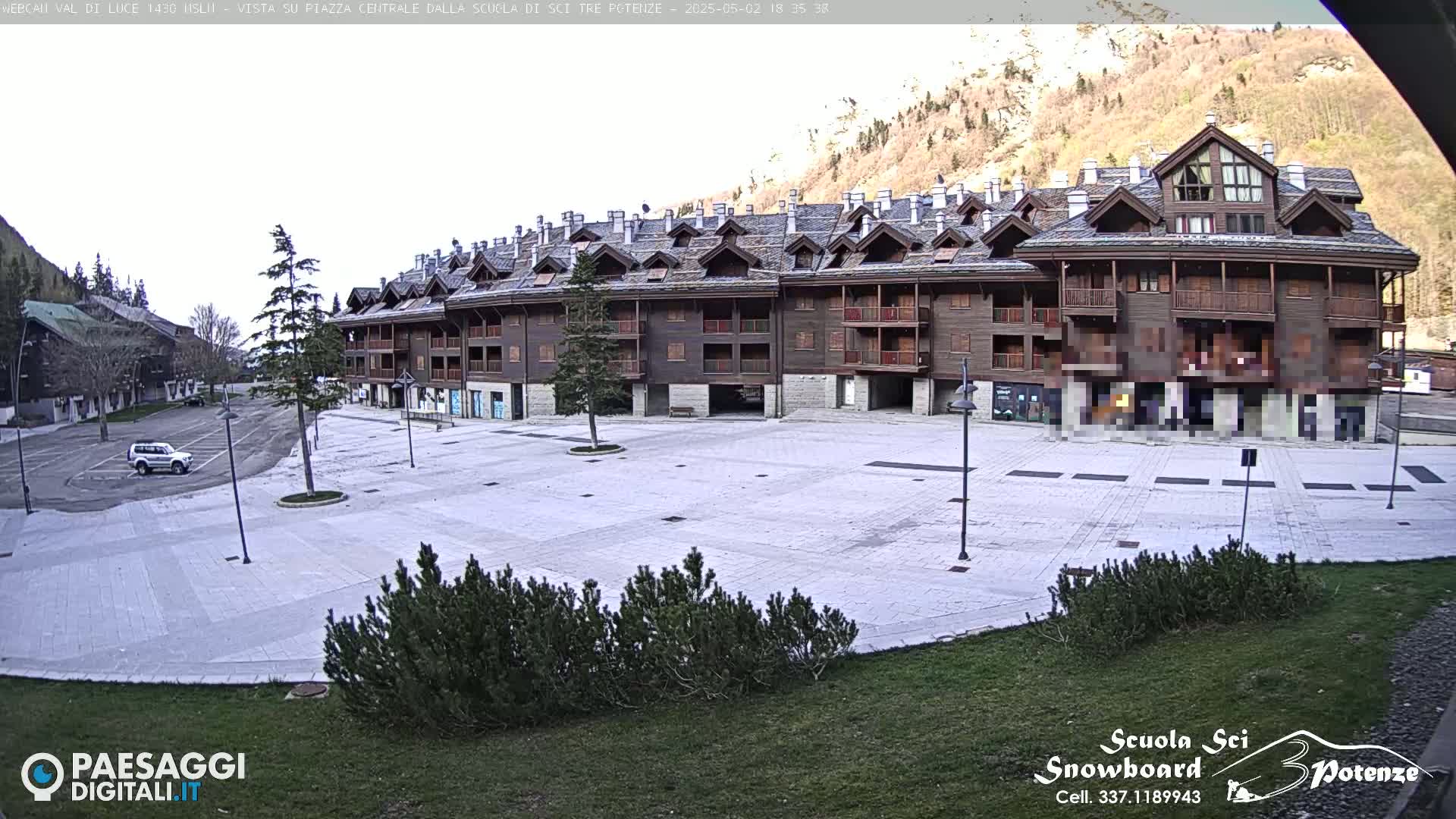 A large, empty plaza is situated in front of a multi-story brown building, with mountains and some trees in the background under a mostly clear sky.