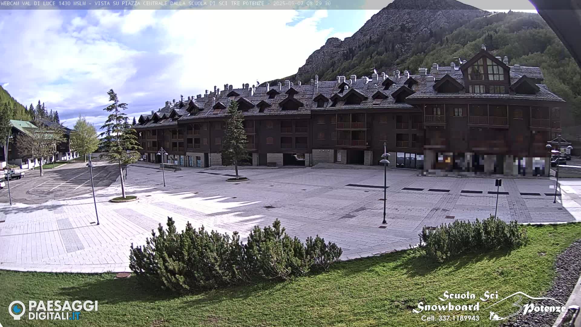A large, empty plaza paved in light-grey stone sits in front of a multi-story brown building, all under a partly cloudy sky in a mountainous region.