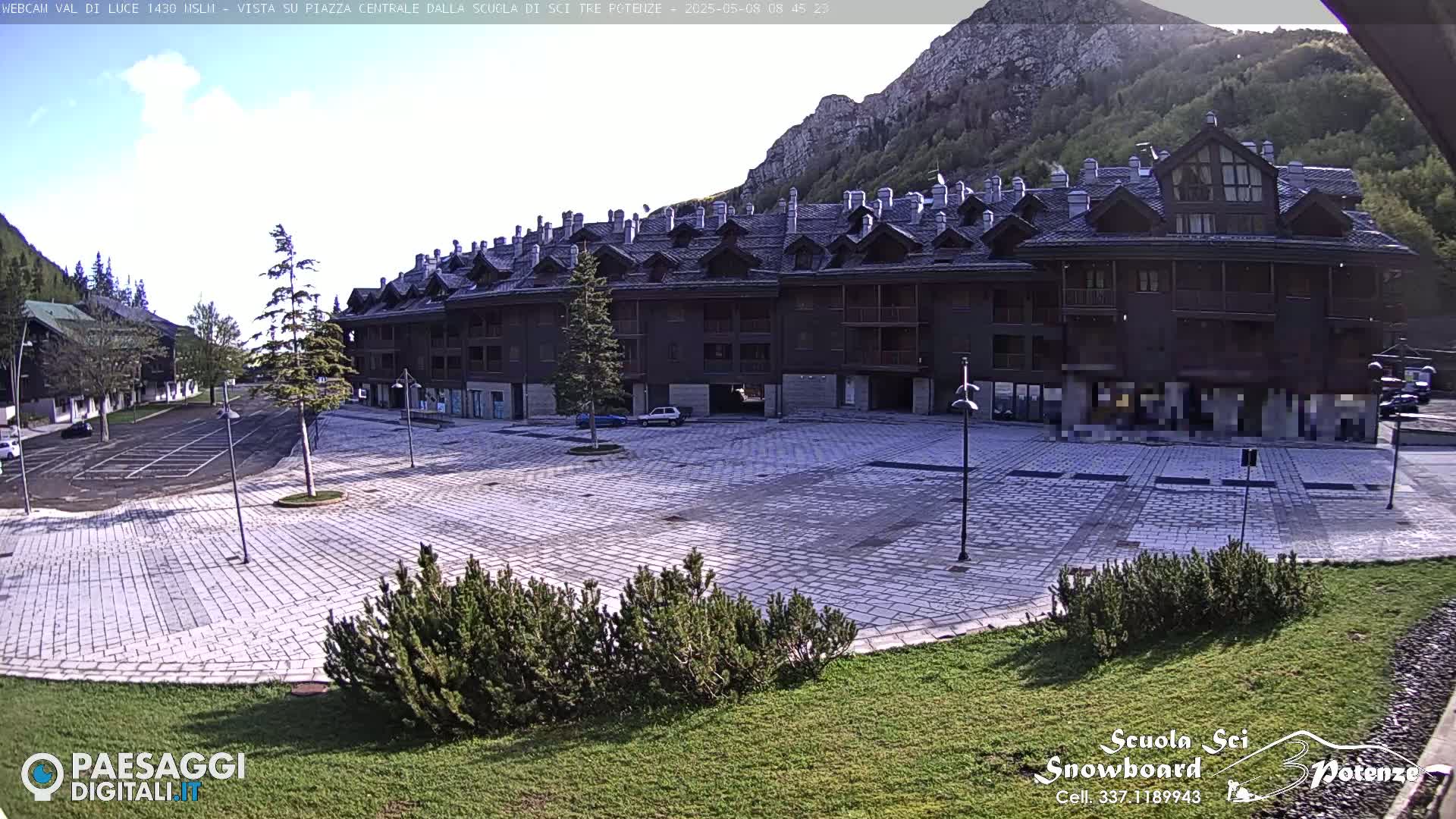 A large, stone paved plaza sits in front of a multi-story building in a mountainous area under a mostly clear sky.