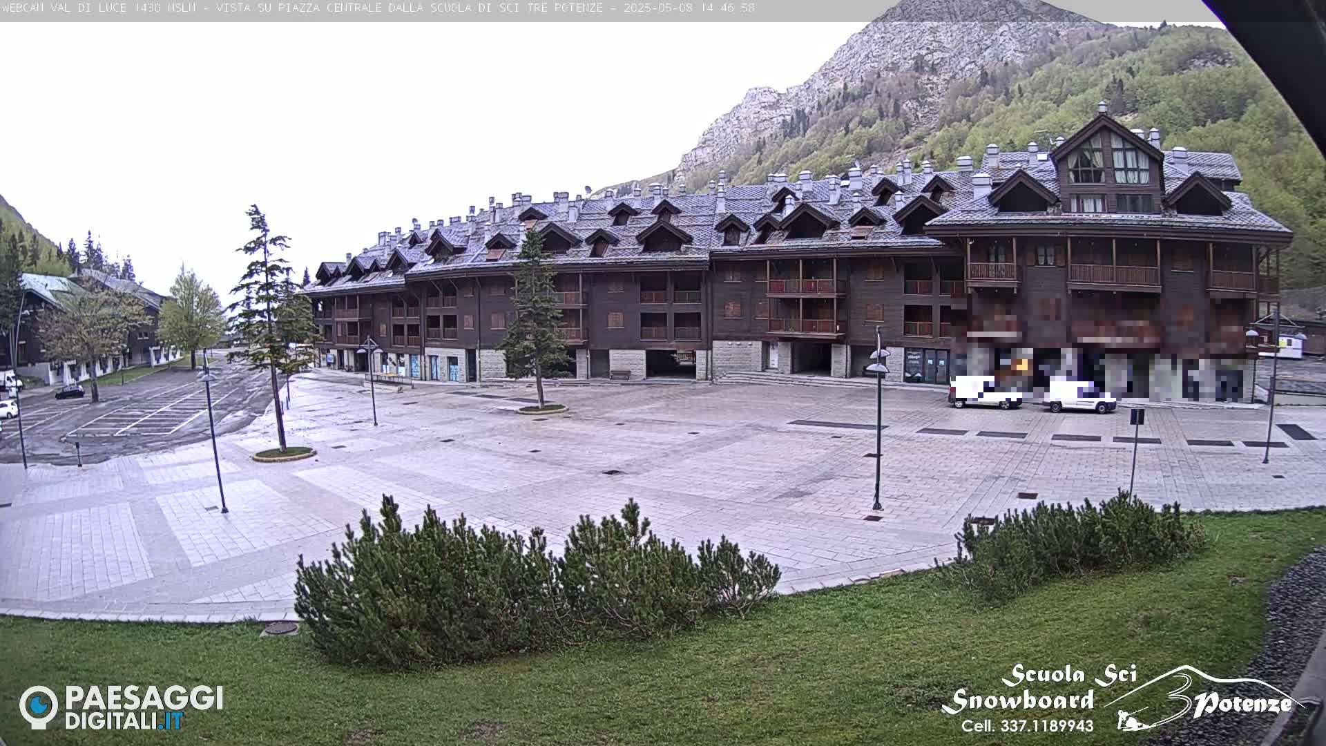 A large, empty plaza sits in front of a multi-story brown building, with mountains and trees visible in the background under an overcast sky.