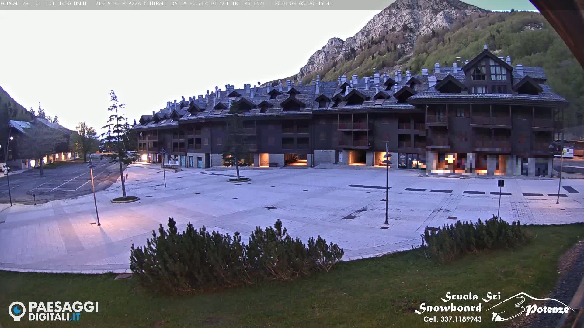A large, empty plaza sits in front of a long, dark-brown building nestled against a mountainside under a clear, twilight sky.