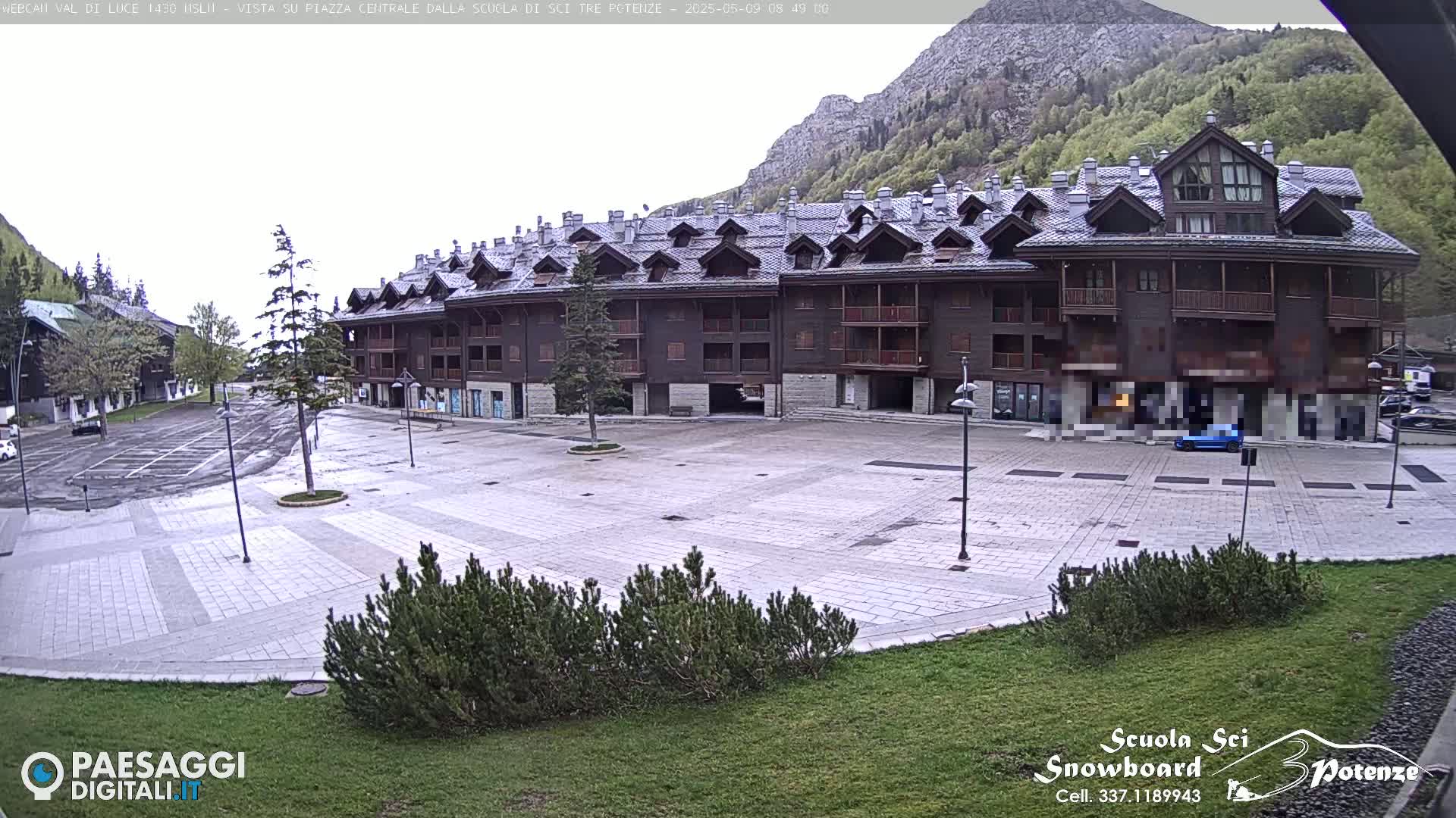 A large, multi-story, dark brown building sits in a paved plaza, surrounded by a few trees and a mountainside in the background under an overcast sky.