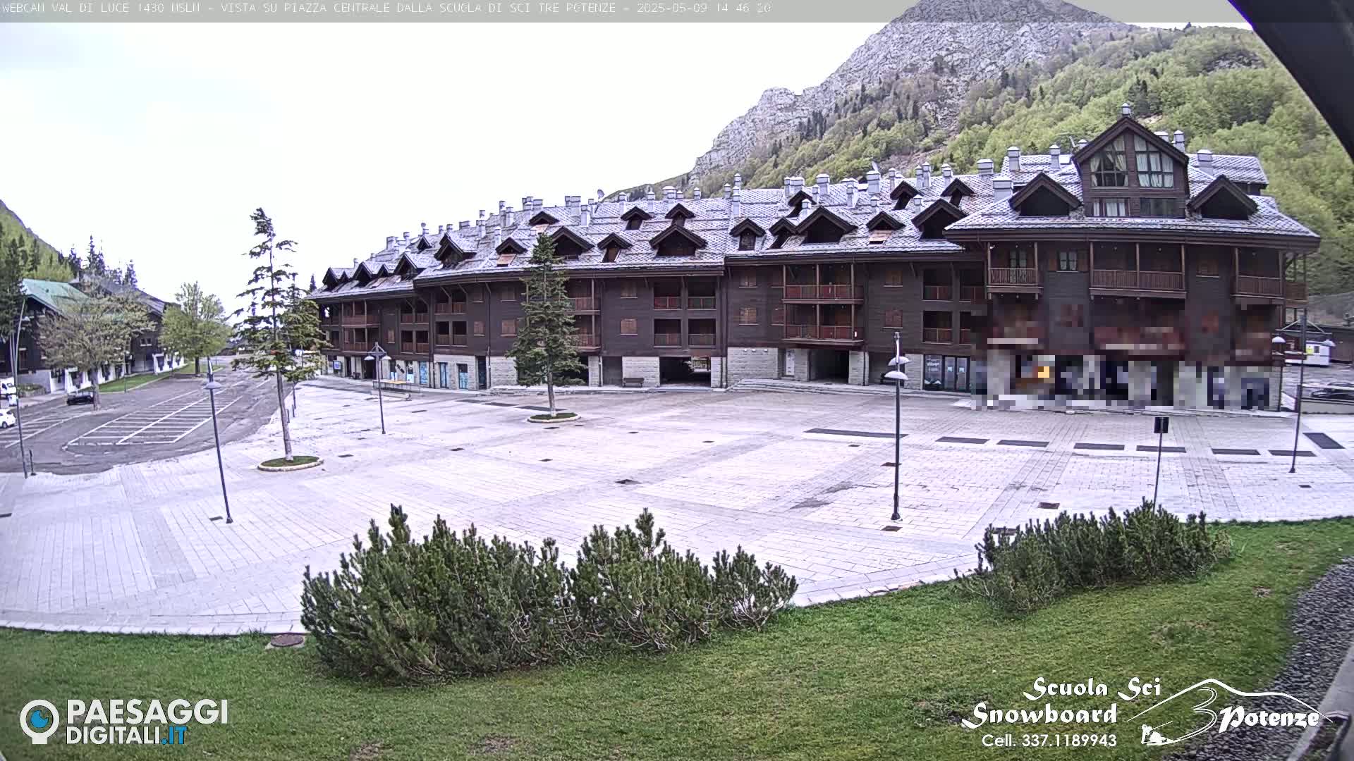 A large, empty plaza sits in front of a multi-story brown building, backed by a mountain range under a mostly clear sky.