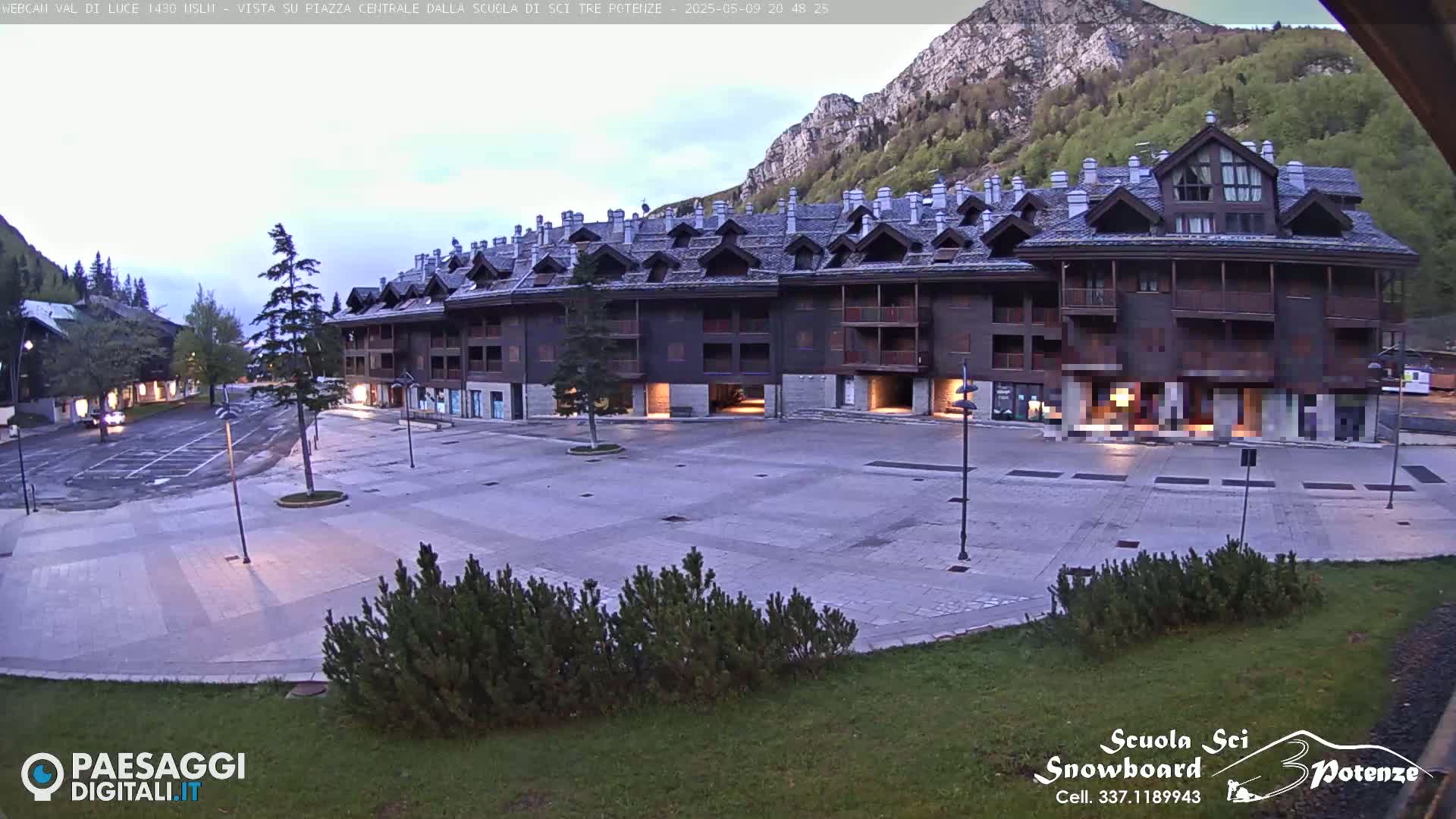 A large, dark-brown building with many windows and a sloped roof sits in a paved plaza next to a mountain under a twilight sky.