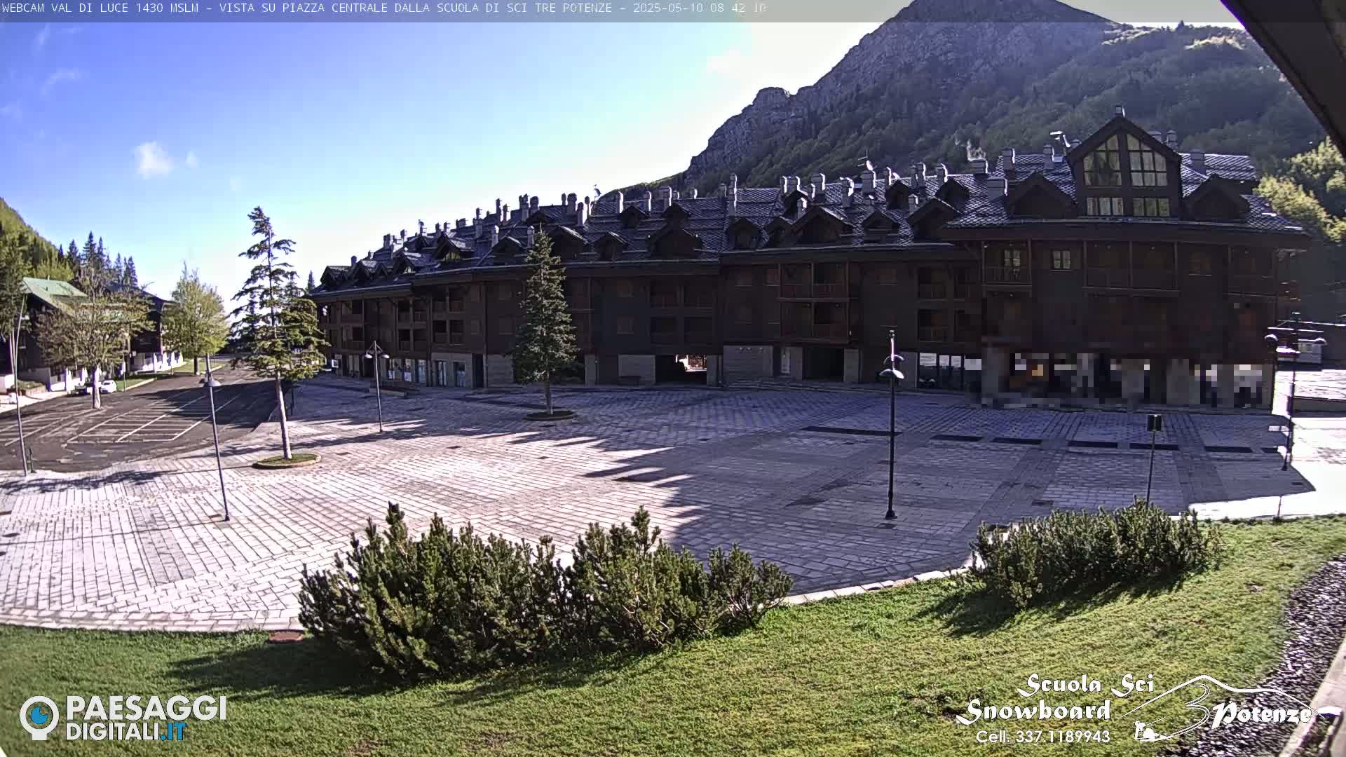 A large, empty paved plaza sits in front of a multi-story brown building, under a mostly sunny sky, with mountains in the background.