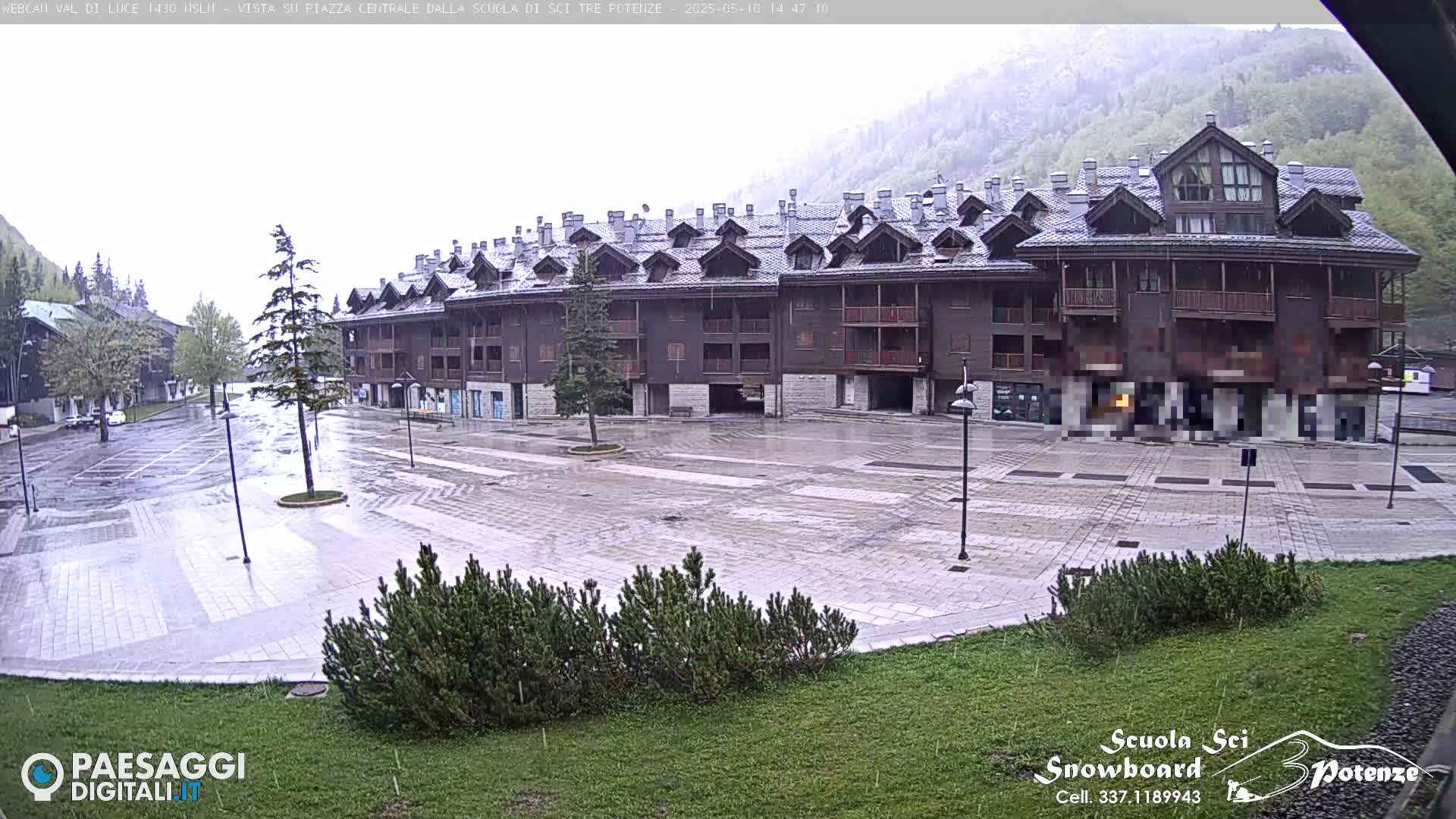 A rain-slicked, empty plaza sits in front of a large, multi-story, dark brown building nestled in a mountain valley.