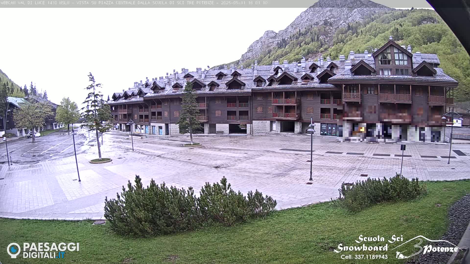 A large, multi-story brown building stands in a mostly empty paved plaza, overlooking a mountainside under an overcast sky.