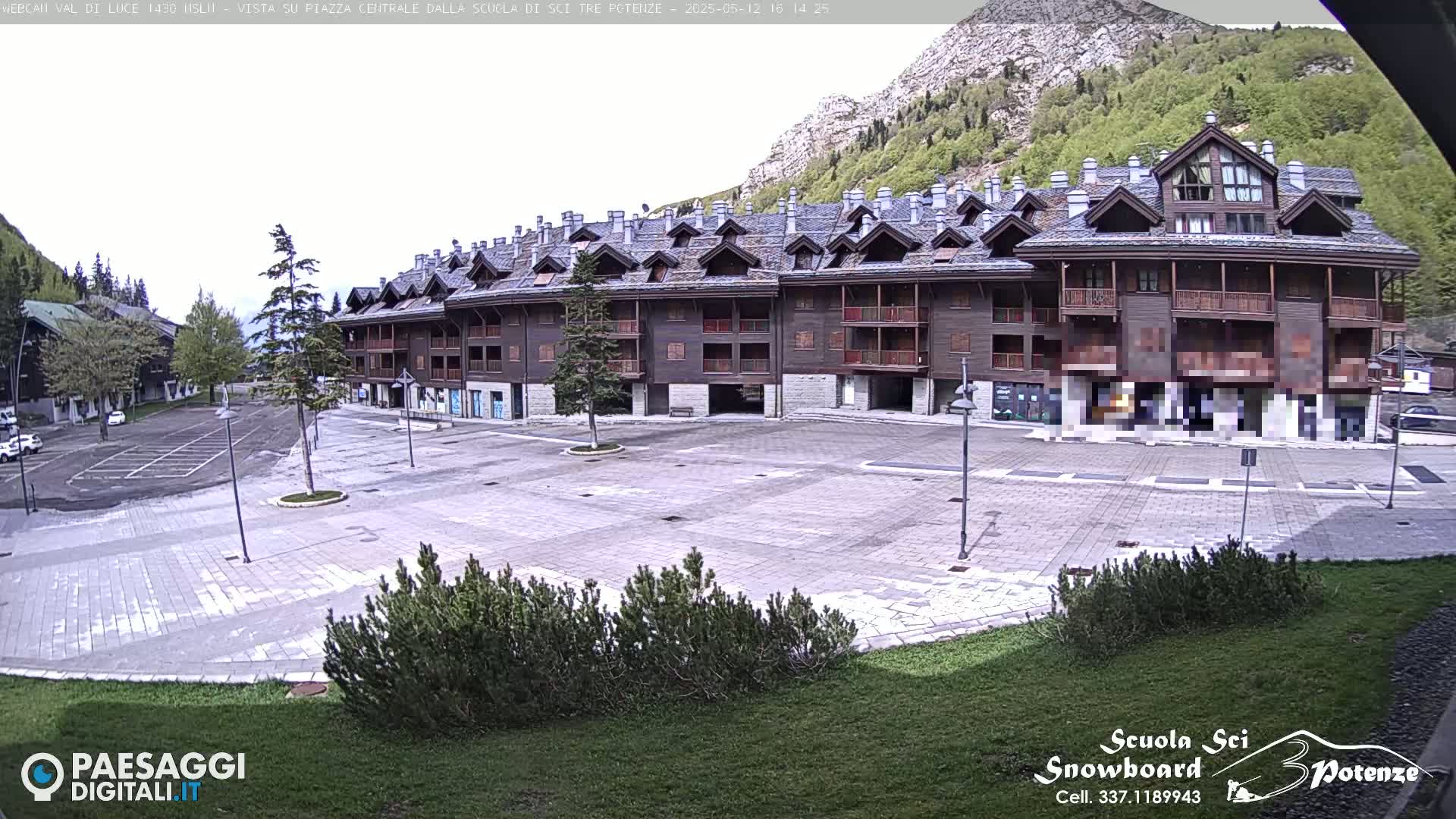 A large, empty paved square sits in front of a multi-story brown building, with a mountain and green trees in the background under an overcast sky.