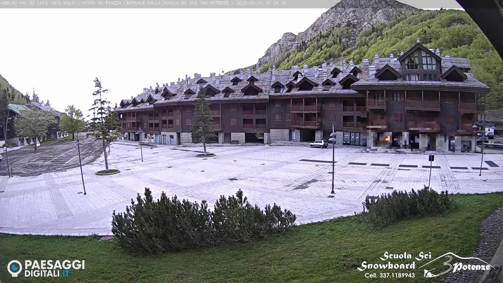 A large, empty paved plaza sits in front of a multi-story brown building, nestled against a mountainside under an overcast sky.