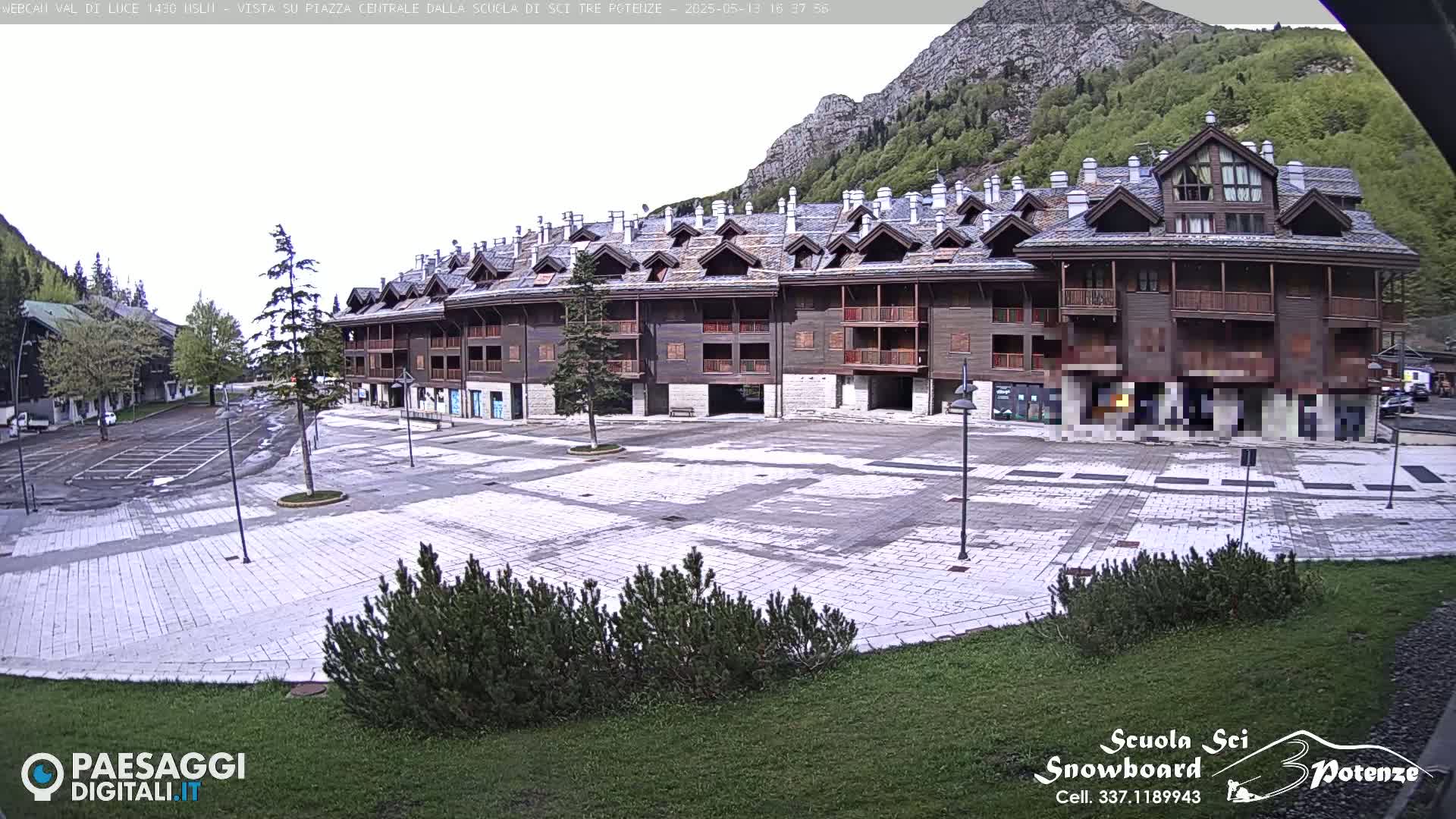 A large, mostly empty plaza is situated in front of a multi-story residential building in a mountainous area under a mostly cloudy sky.