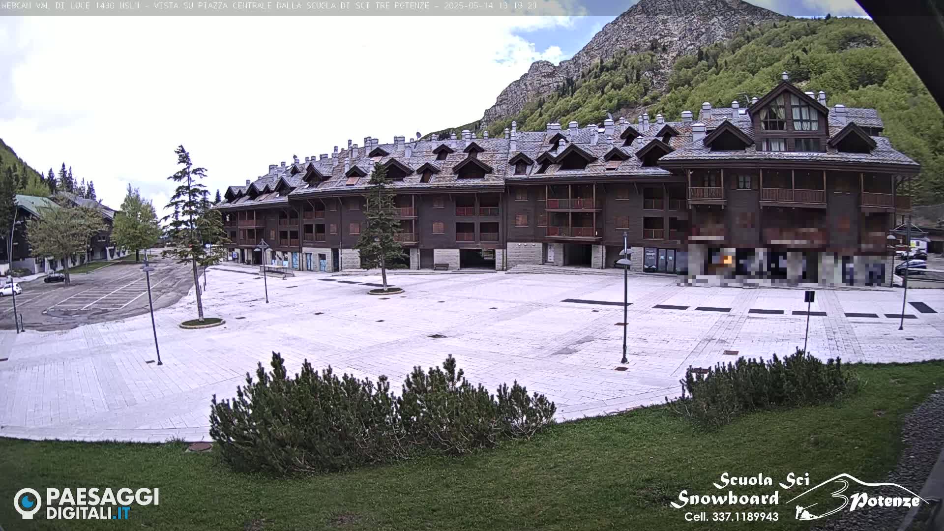 A large, empty plaza paved with light-colored stone is situated in front of a multi-story brown building, with mountains and greenery visible in the background under a partly cloudy sky.