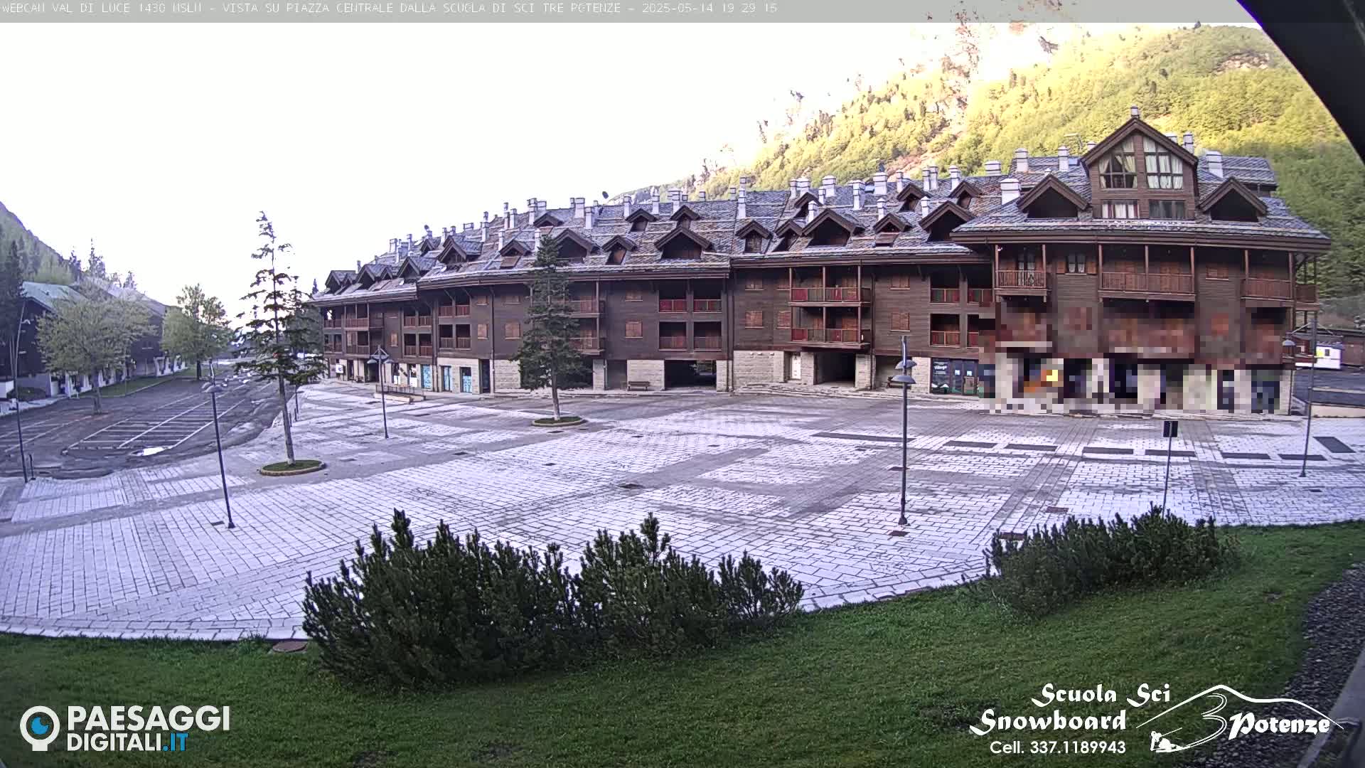 A large, stone plaza sits in front of a multi-story, wood-sided building complex, nestled against a mountainside under a bright, overcast sky.