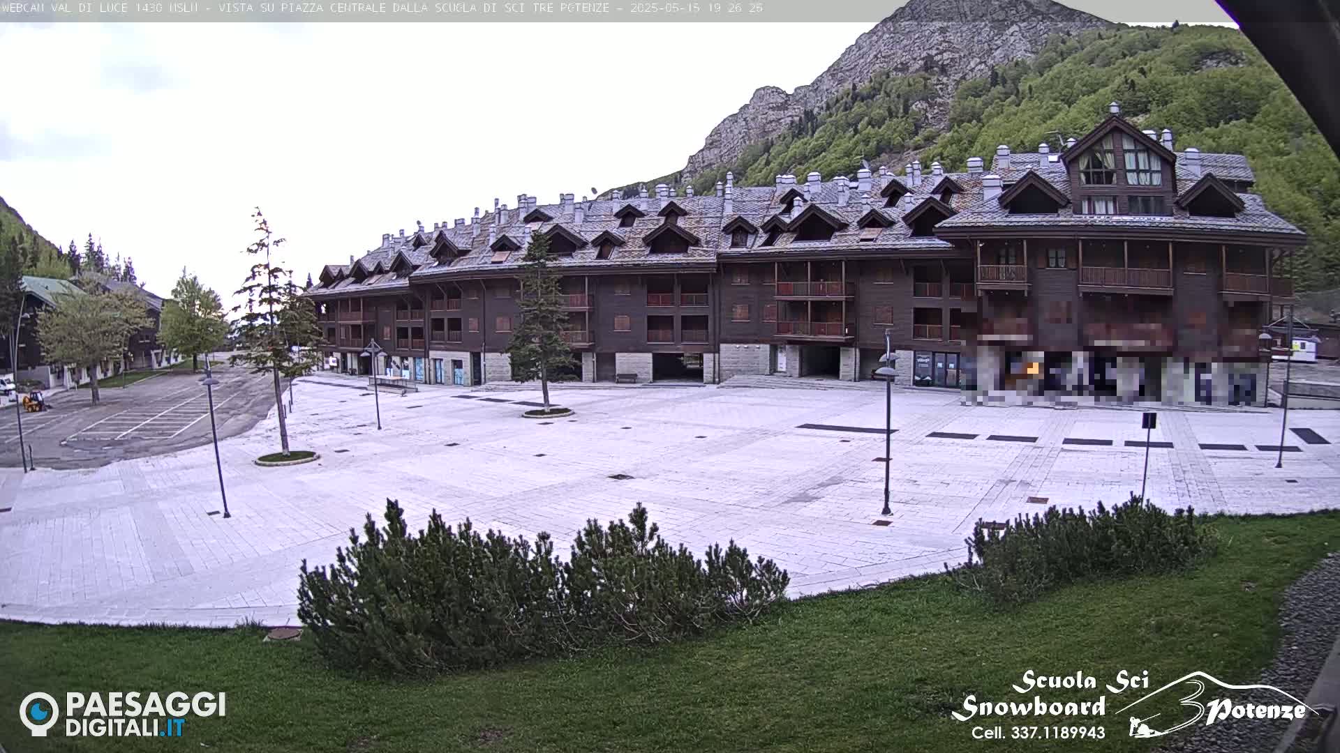 A large, empty plaza paved in light-colored stone is situated in front of a multi-story brown building against a backdrop of mountains under an overcast sky.