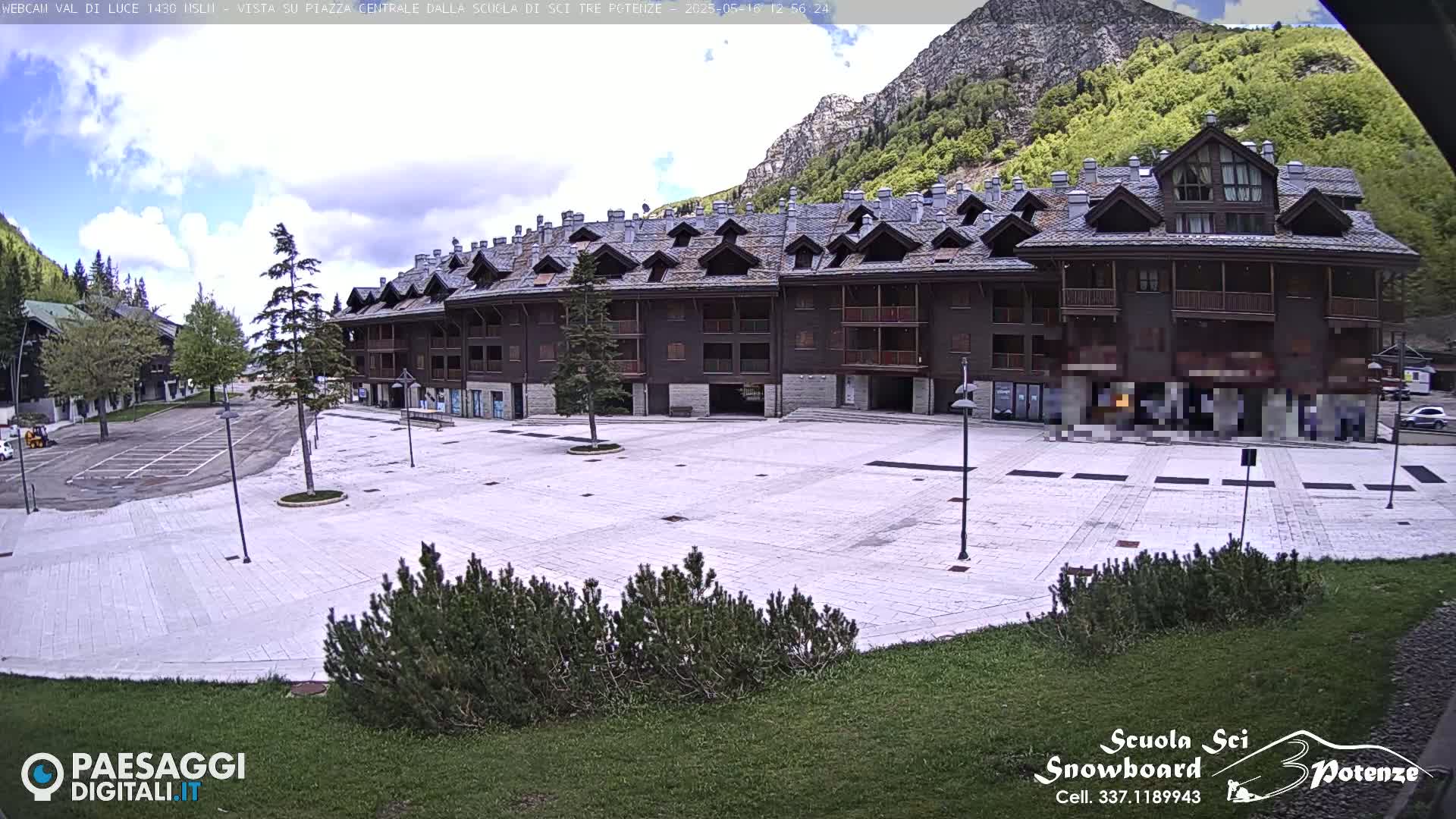 A large, empty plaza paved in light-colored stone sits in front of a multi-story brown building, all under a partly cloudy sky with mountains in the background.