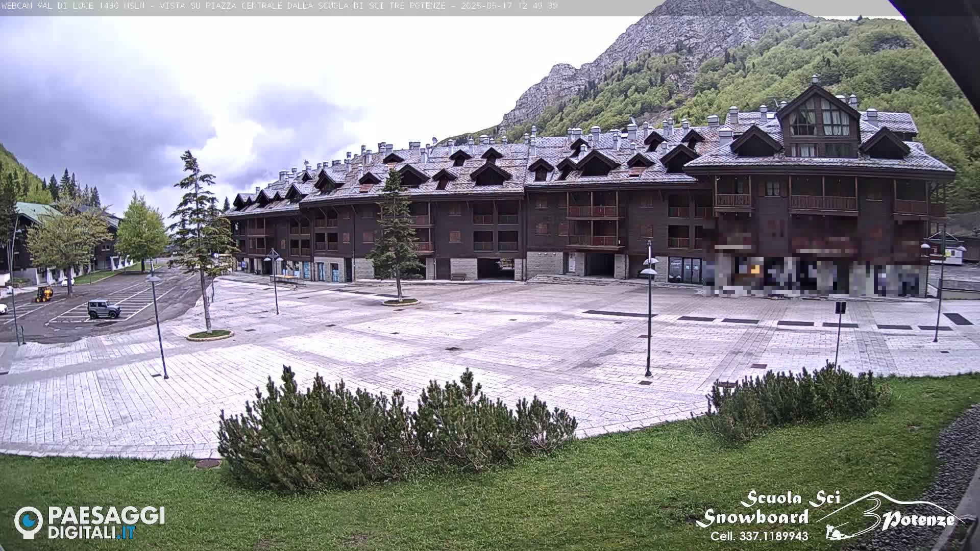 A large, light-grey paved plaza sits in front of a multi-story, dark-brown building, with a mountain and cloudy sky visible in the background.