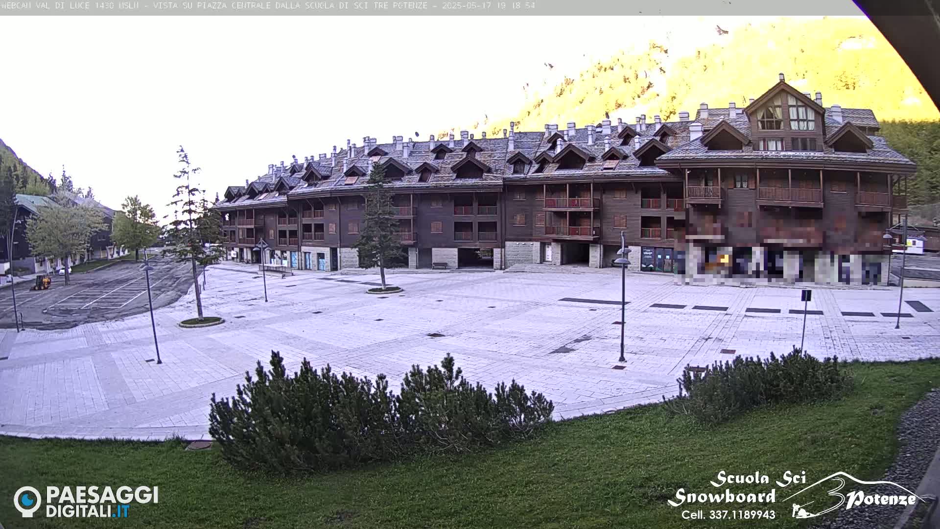 A large, empty paved plaza sits in front of a multi-story wooden apartment building, with mountains and a mostly clear sky in the background.