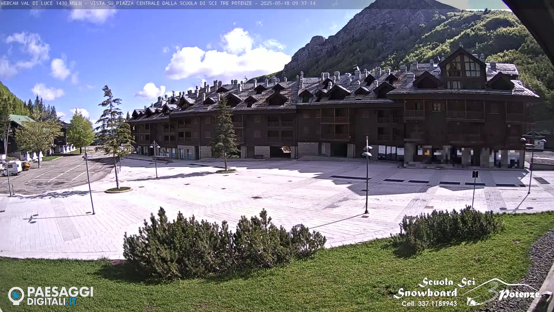A mostly empty paved plaza sits in front of a large multi-story building, with mountains visible in the background under a partly cloudy sky.