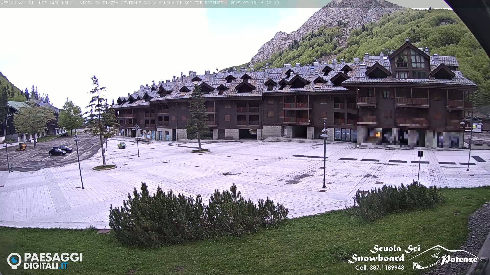 A large, empty paved square sits in front of a multi-story, dark-brown building against a backdrop of a mountain and overcast sky.