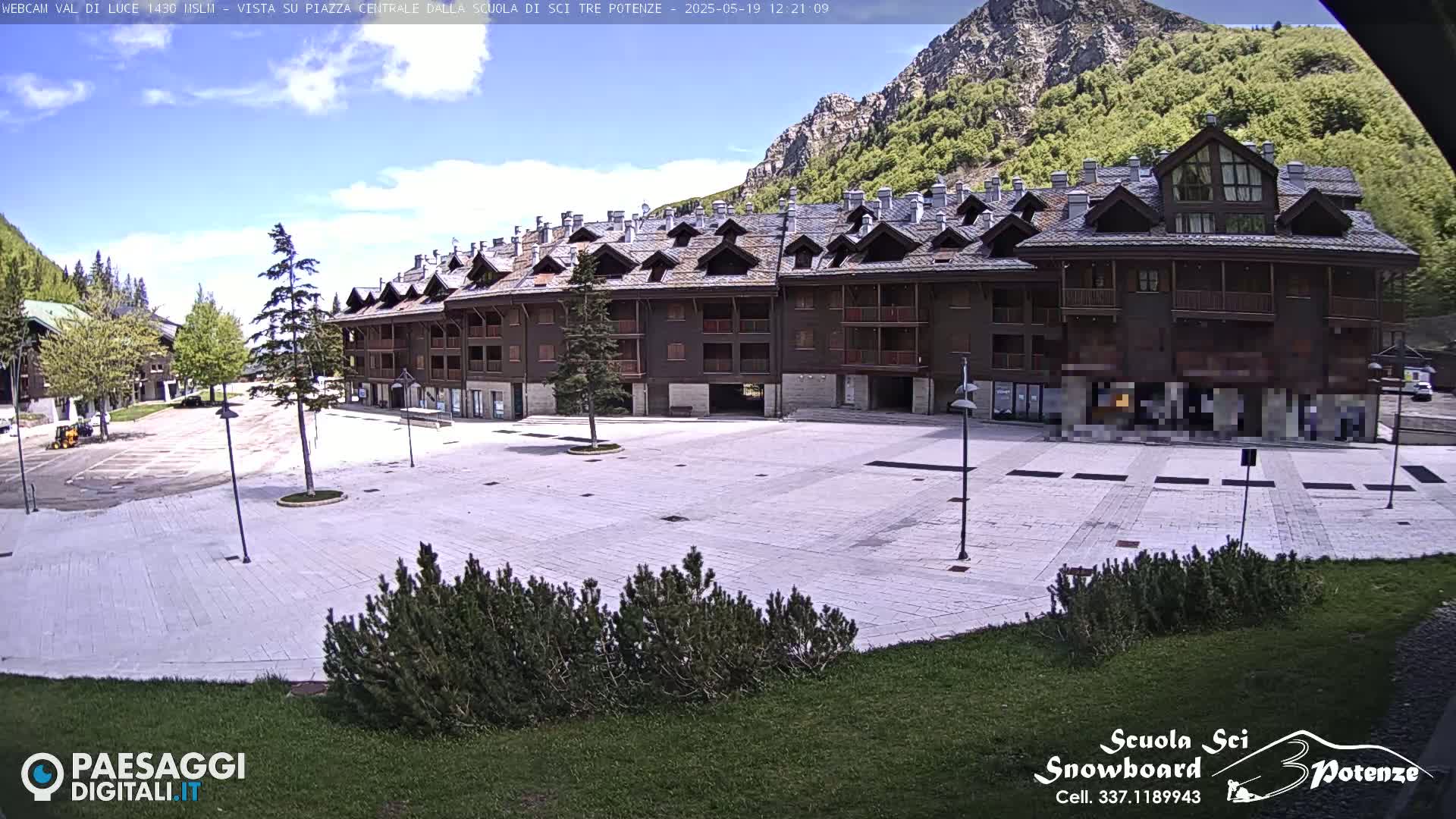 A large, empty paved square sits in front of a multi-story brown building, with mountains and green trees in the background under a partly cloudy sky.