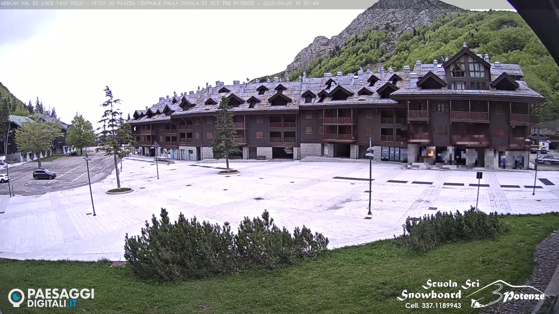 A large, mostly empty plaza paved with light-colored stones sits in front of a multi-story, dark brown building against a backdrop of mountains under an overcast sky.