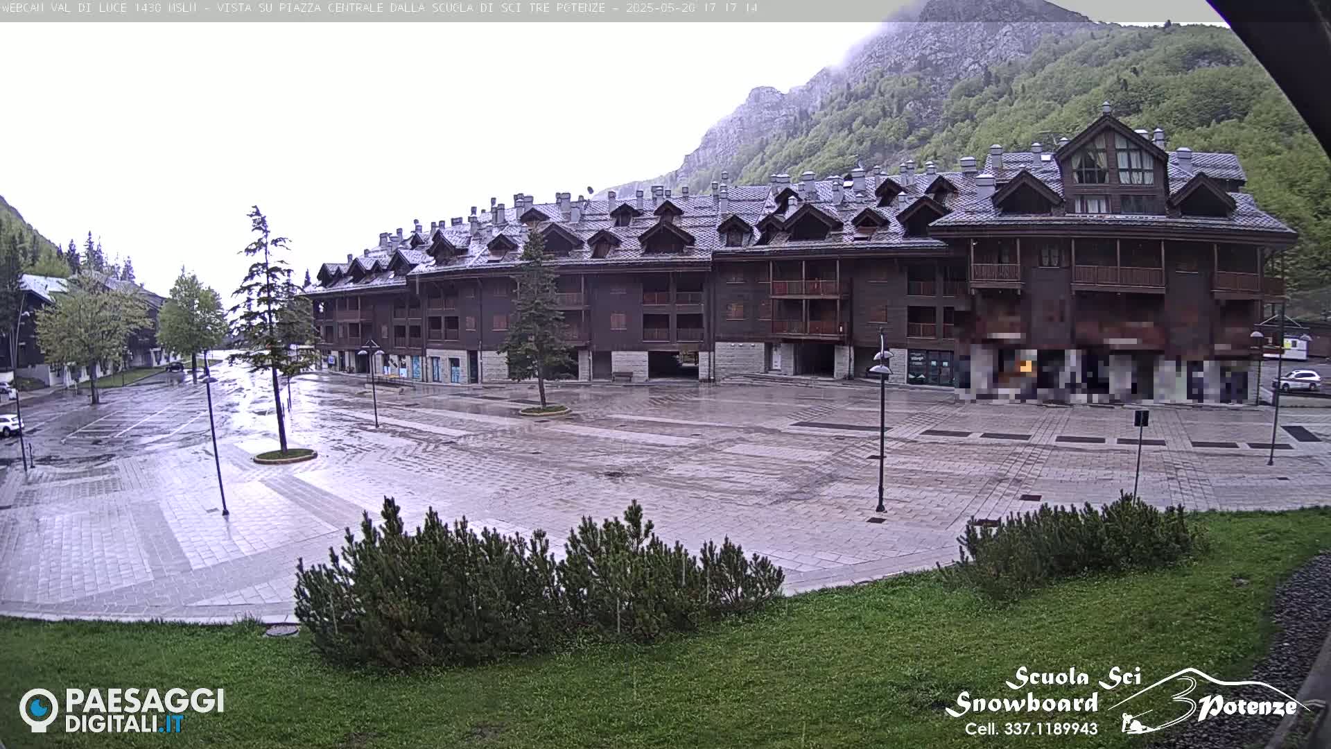 A large, dark-brown building sits across a wet, paved plaza under an overcast sky, with mountains visible in the background.