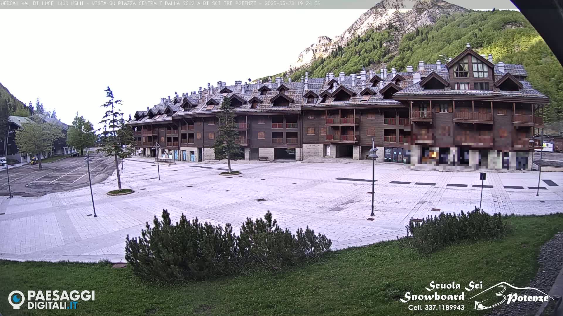 A large, empty paved plaza sits in front of a multi-story brown building, with mountains and trees visible in the background under a bright, mostly clear sky.