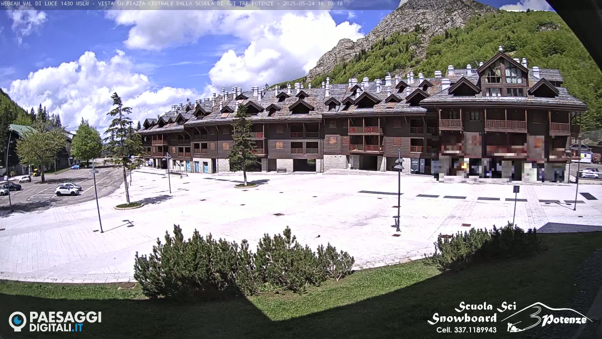 A large, light-colored plaza sits in front of a long, multi-story, wood-sided building, all under a partly cloudy sky, with a mountain visible in the background.