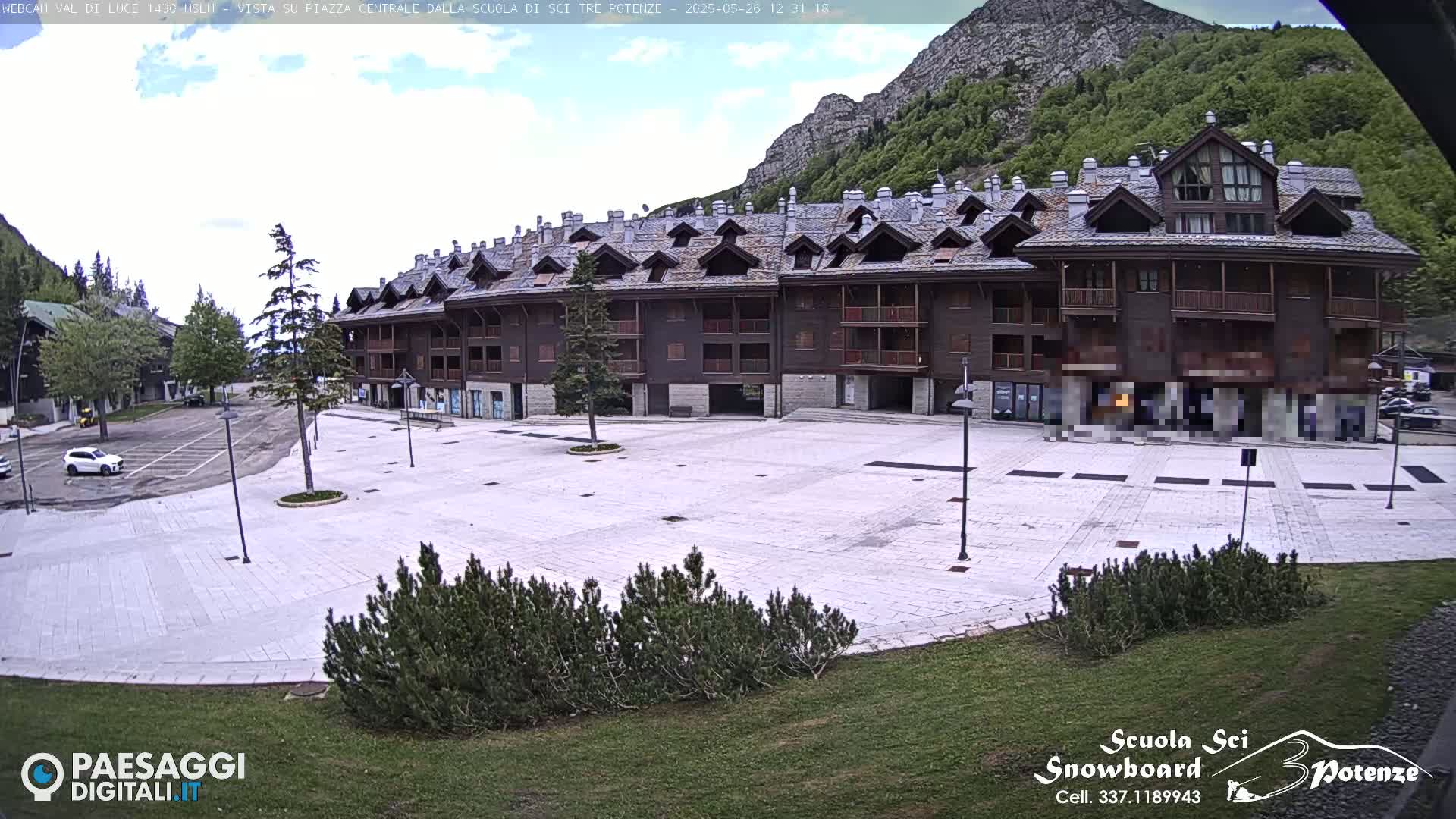A large, empty plaza is in front of a multi-story, wood-sided building, with mountains and greenery visible in the background under a partly cloudy sky.