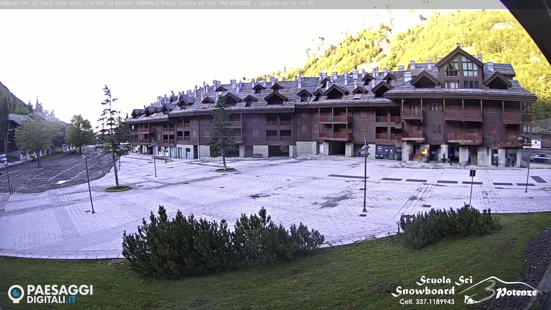 A large, empty paved plaza sits in front of a multi-story wood and stone building, with mountains and trees in the background under a mostly cloudy sky.