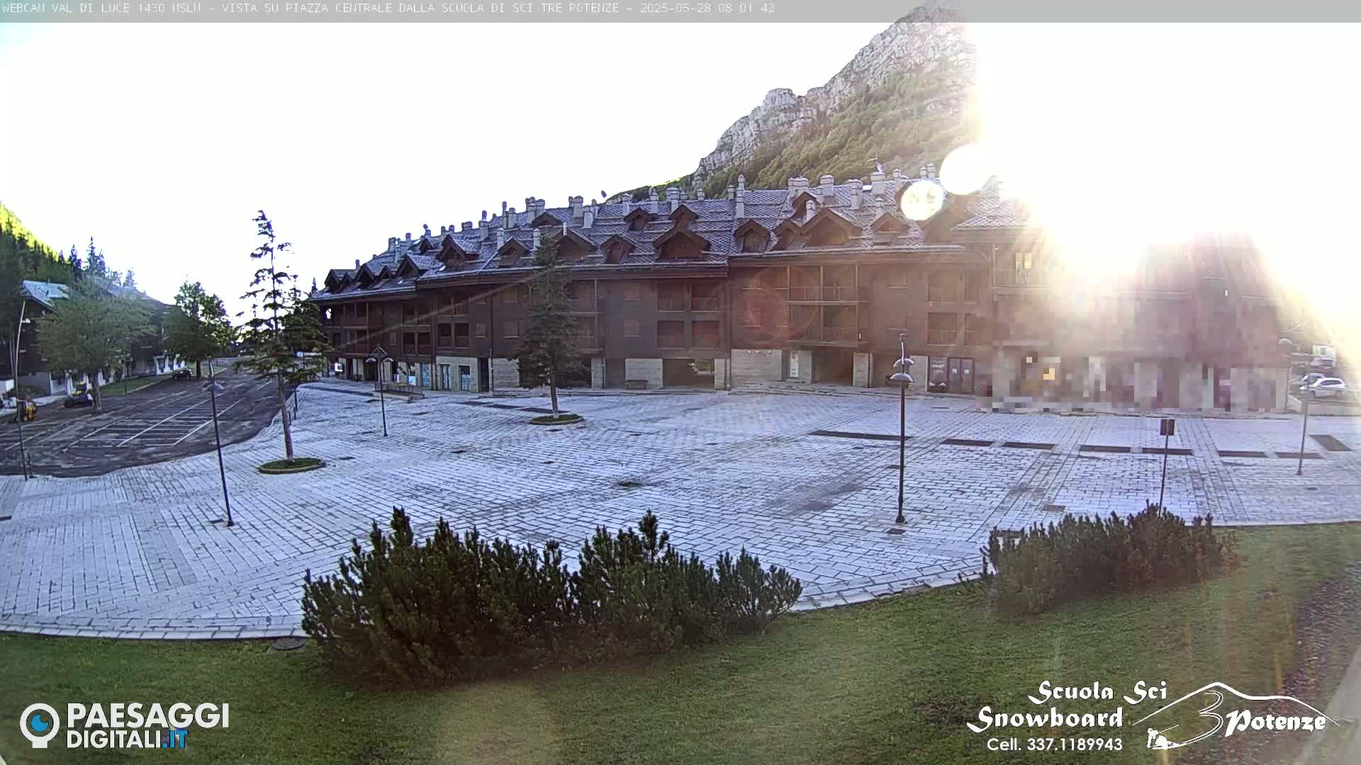 A sunlit, paved plaza sits in front of a large, multi-story brown building, with mountains and trees in the background.