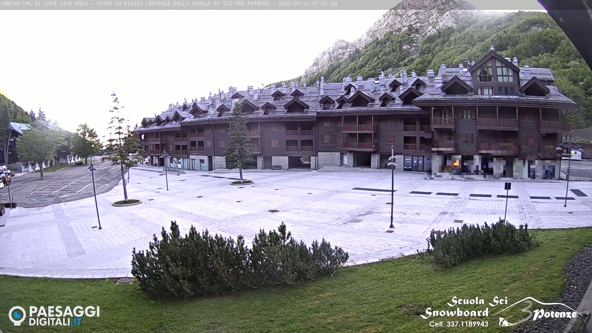 A large, empty paved plaza sits in front of a multi-story brown building, all situated against a backdrop of a mountainside covered in green trees under an overcast sky.