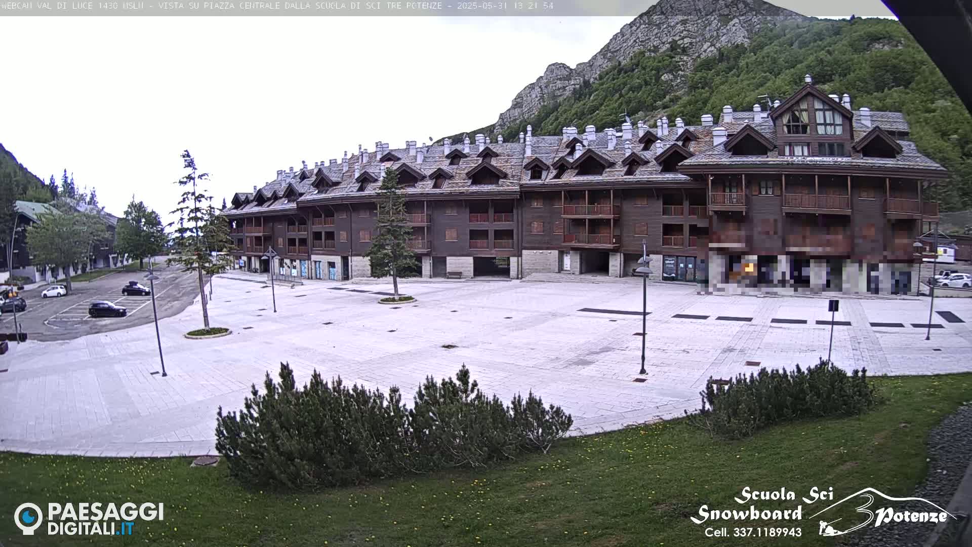 A large, empty plaza in front of a multi-story building, situated against a backdrop of mountains under an overcast sky.