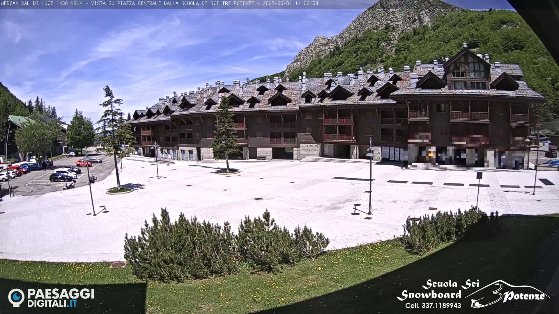 A large, light-colored plaza sits in front of a multi-story brown building, with mountains and a sunny sky in the background.