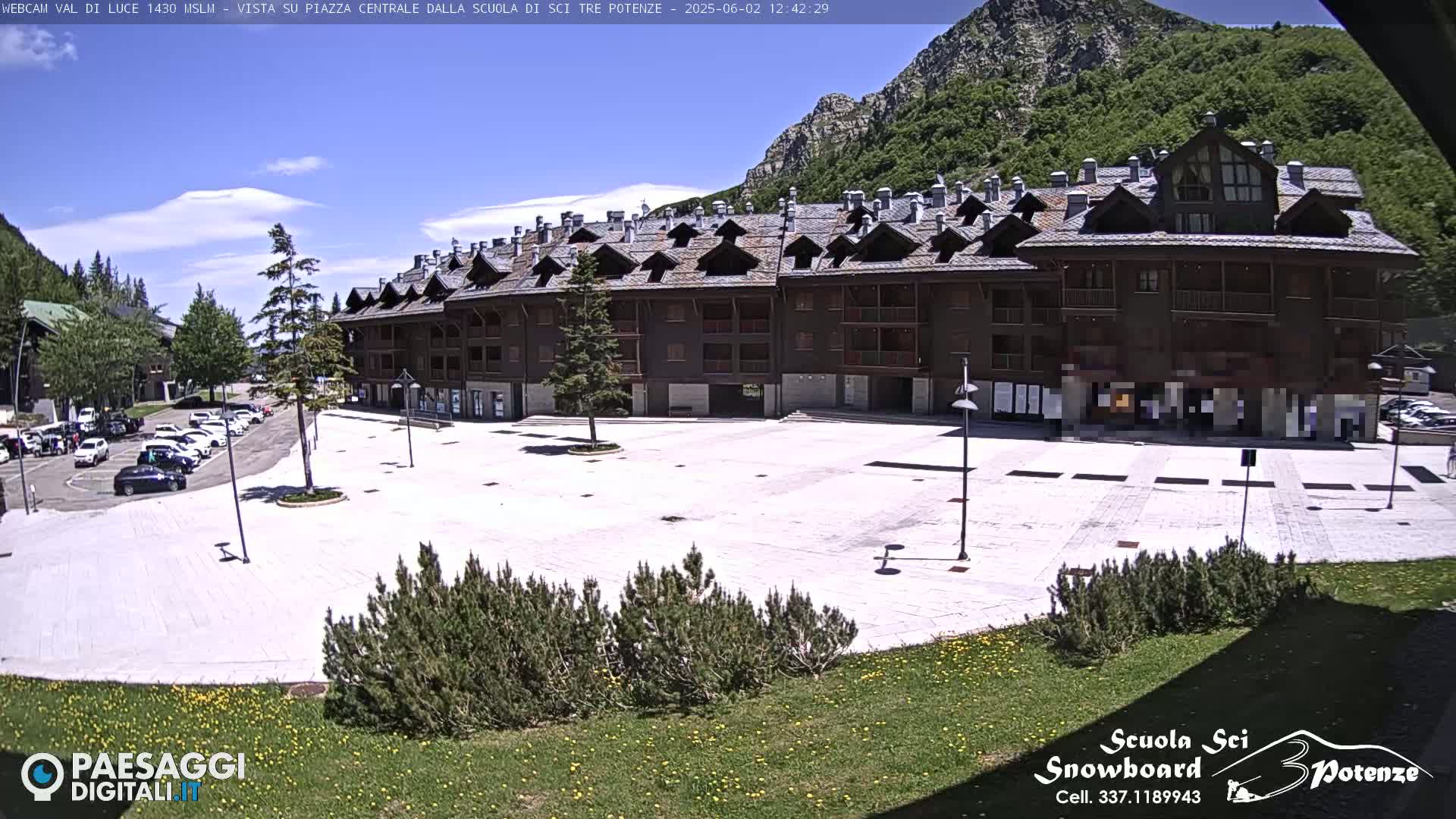 A mostly empty paved plaza sits in front of a large, multi-story brown building, with mountains and a clear blue sky visible in the background.