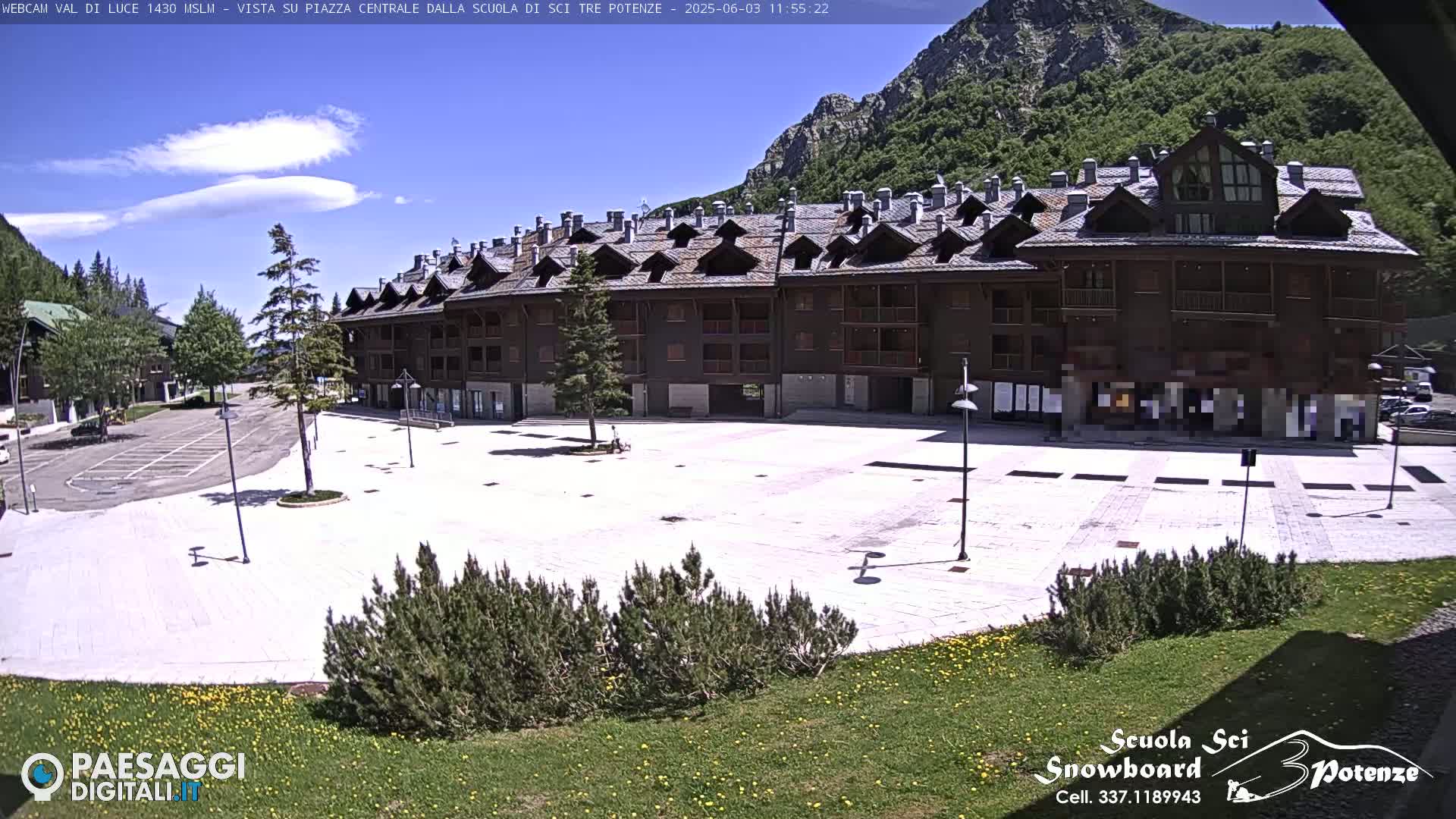 A large, empty plaza paved in light-colored stone is situated in front of a multi-story brown building, with mountains and a partly cloudy blue sky in the background.