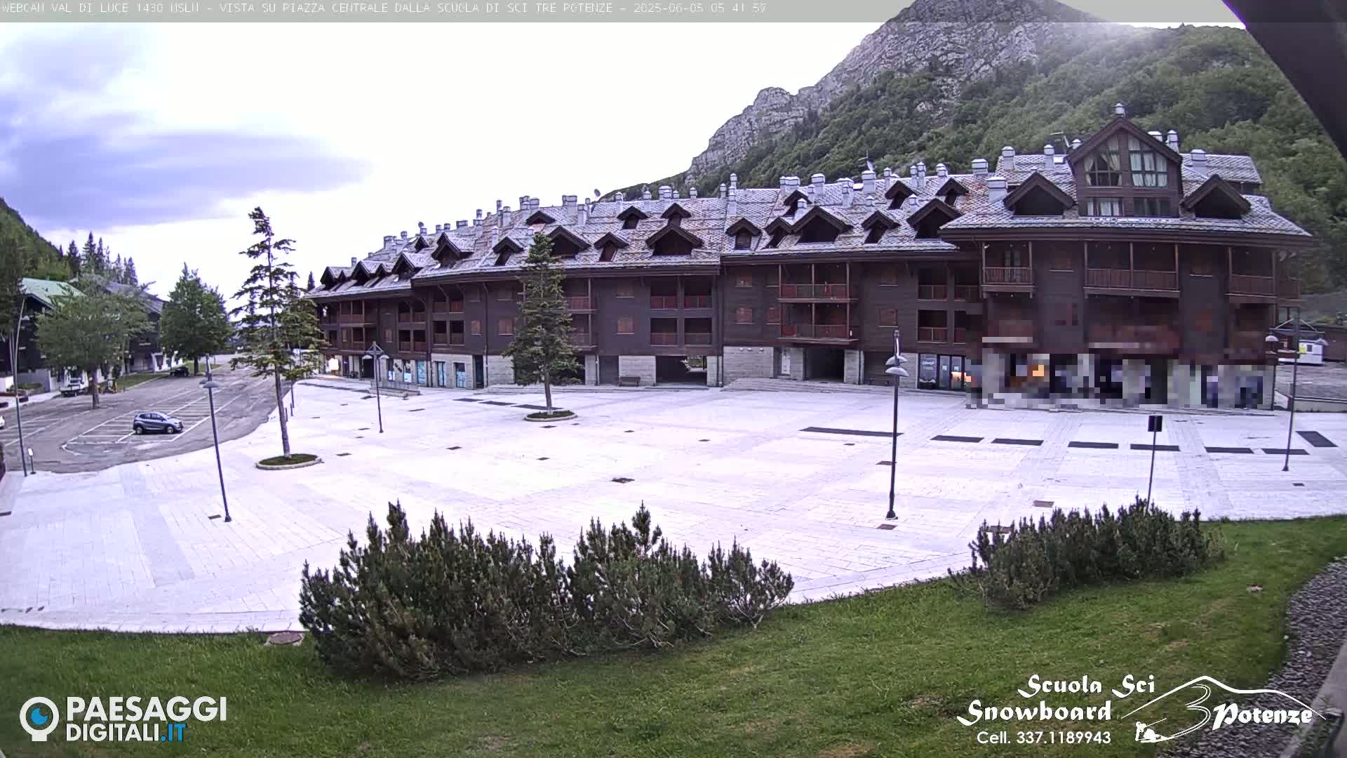 A large, light-colored paved plaza sits in front of a multi-story brown building, with a mountain and overcast sky in the background.